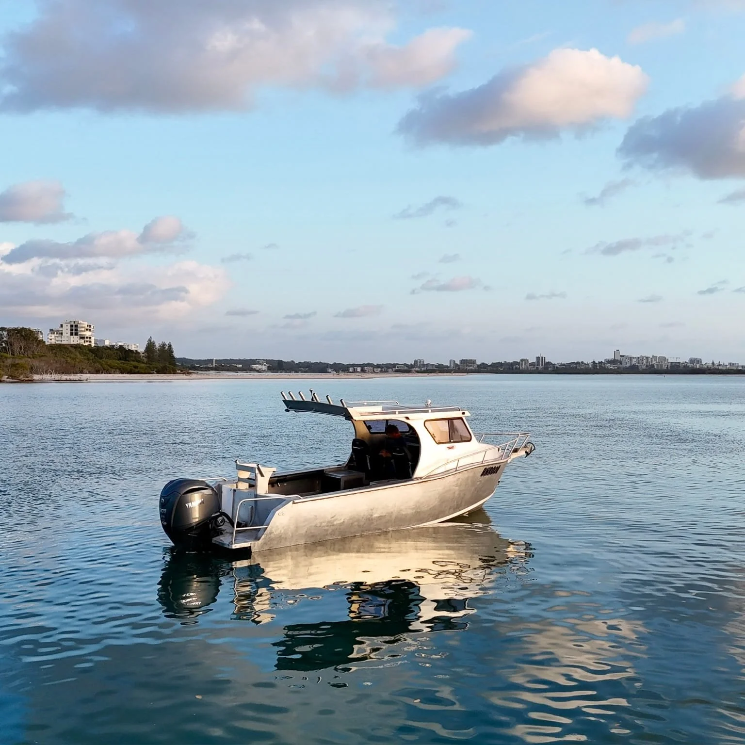 A small white motorboat floating on calm water with a city skyline and partly cloudy sky in the background