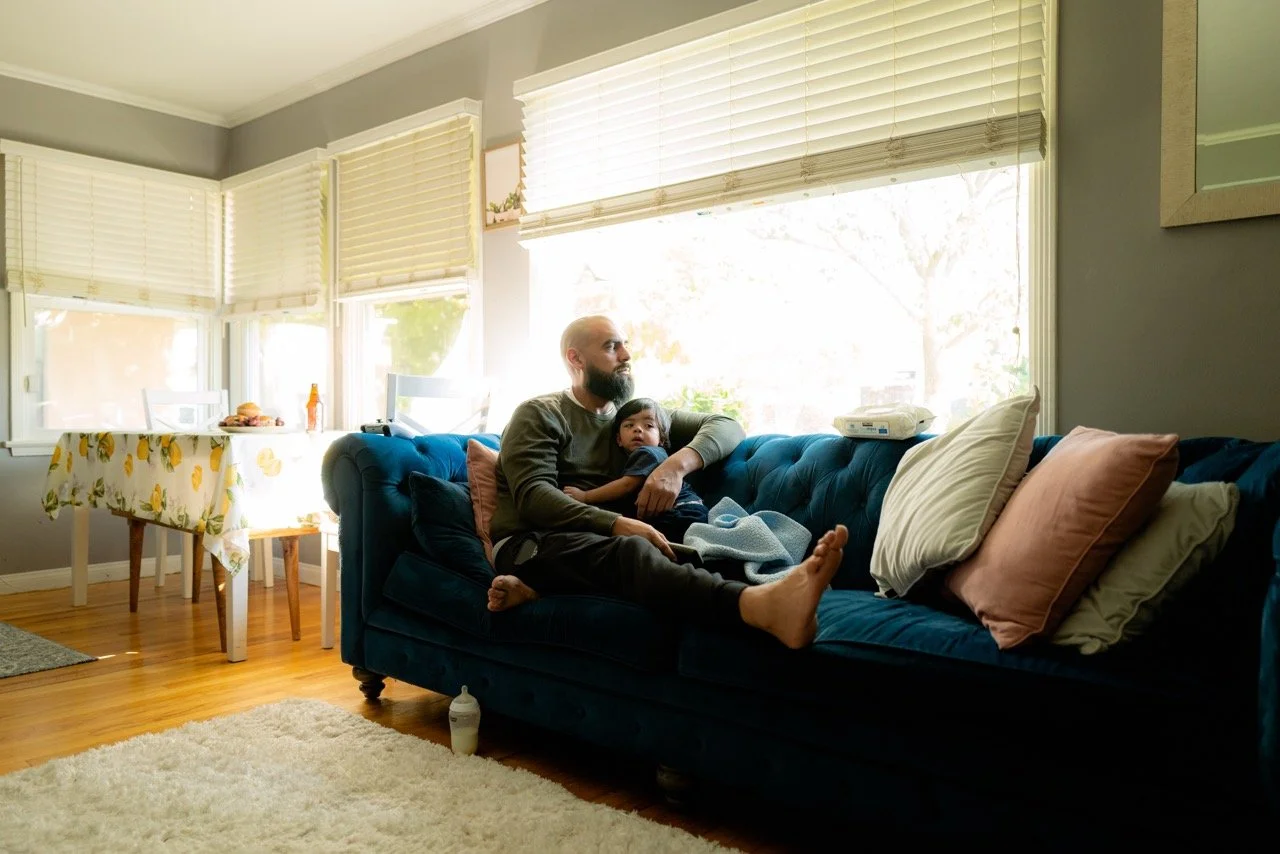 A man with a beard and a young girl sitting on a dark blue couch in a living room, with large windows behind them letting in sunlight, and pillows and a baby bottle on the floor.