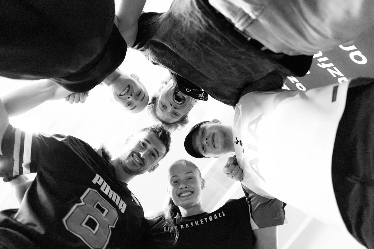 Group of people in a huddle, smiling and looking down at the camera, taken from below in black and white.