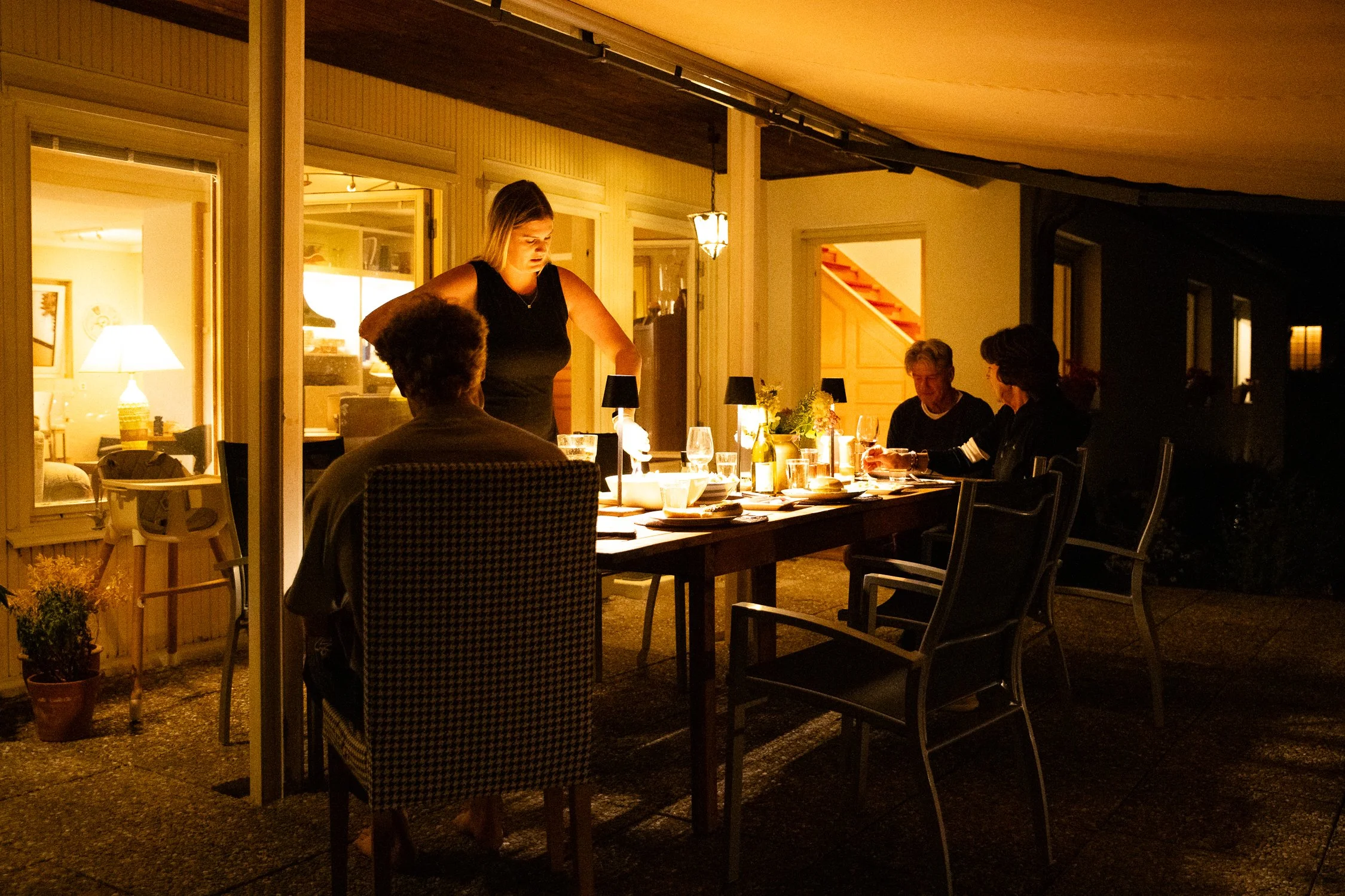 A woman serving food at a dinner table outside a house during the evening, with three people sitting at the table and a warm light from inside illuminating the scene.