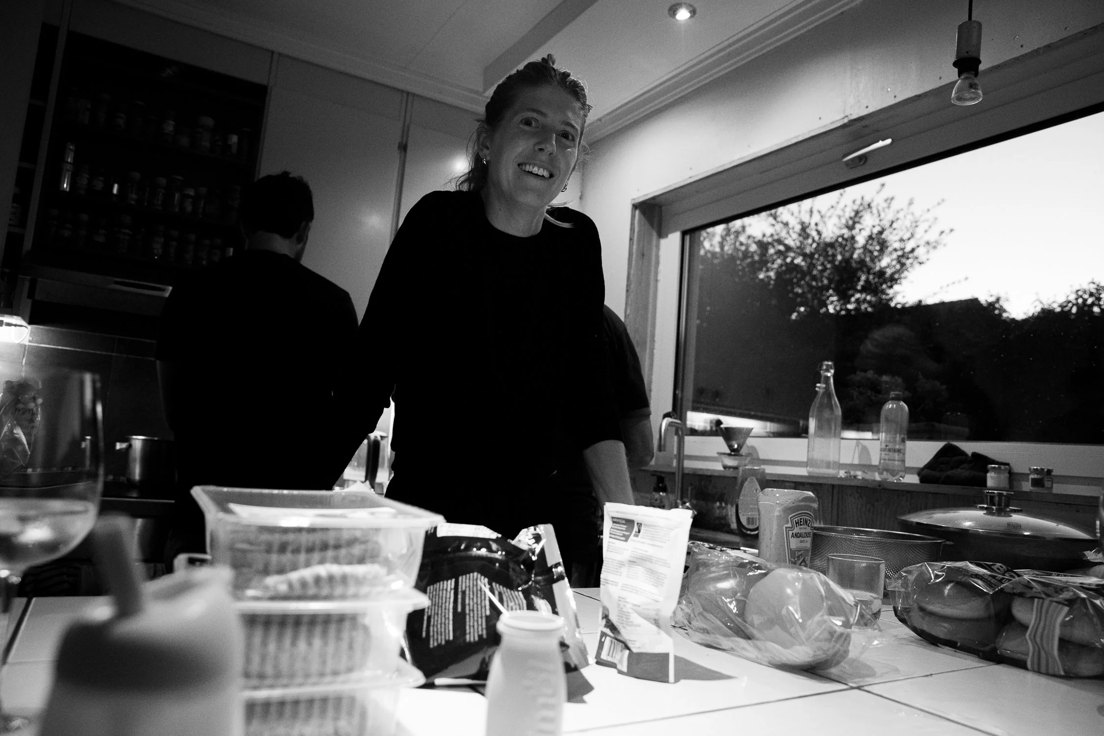 A woman with light skin and long hair smiling in a kitchen, surrounded by various food items and kitchen utensils. Behind her is a large window showing trees outside. The kitchen has cabinets, shelves with jars, and hanging light bulbs.