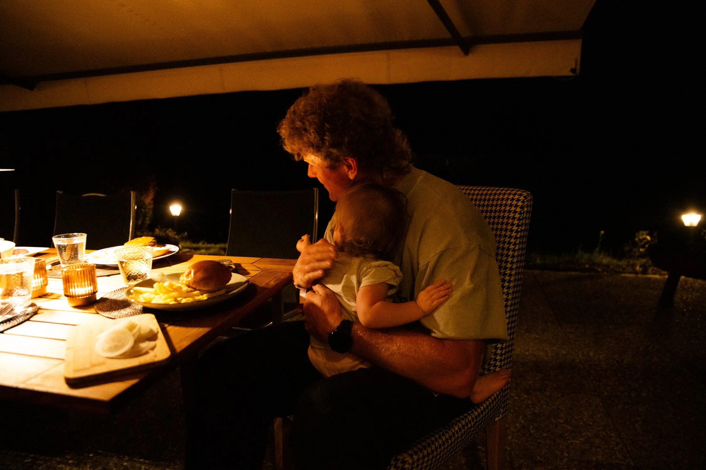 An older person and a child sharing a hug at a dinner table outdoors at night, with lit candles and food on the table.