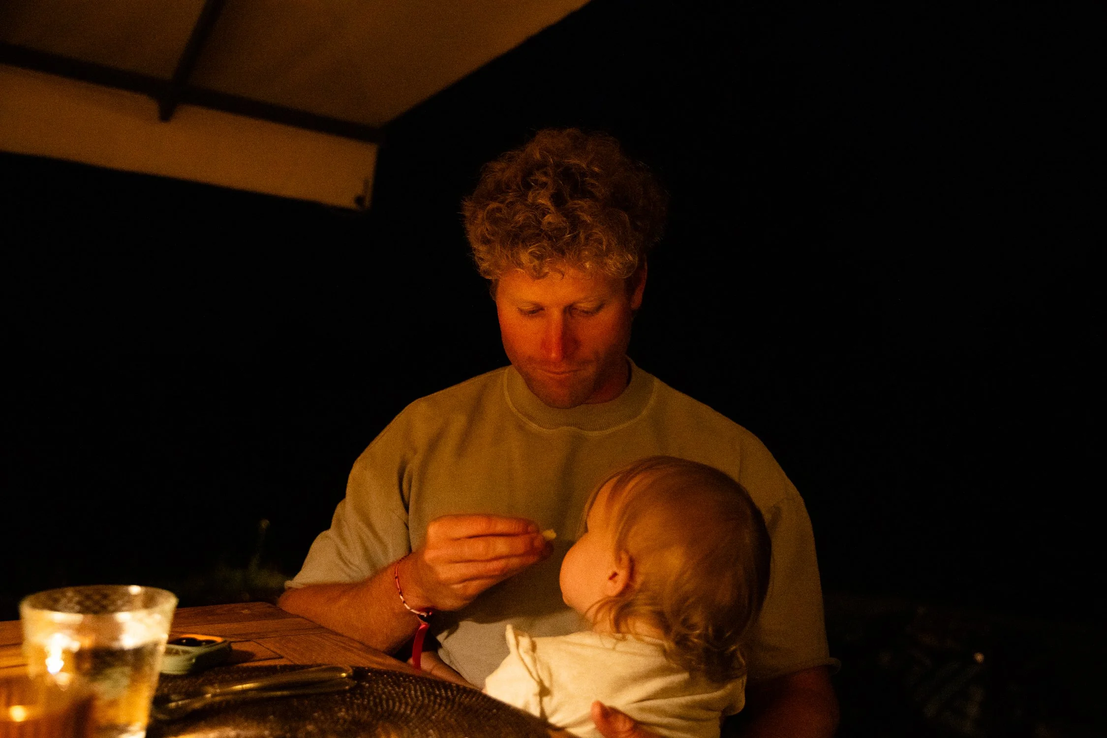 A man with curly blond hair and a light-colored shirt is sitting at a table, looking down at a small child with wavy blond hair, who is facing away from the camera. The scene is dimly lit with warm lighting, and the background is dark and out of focu