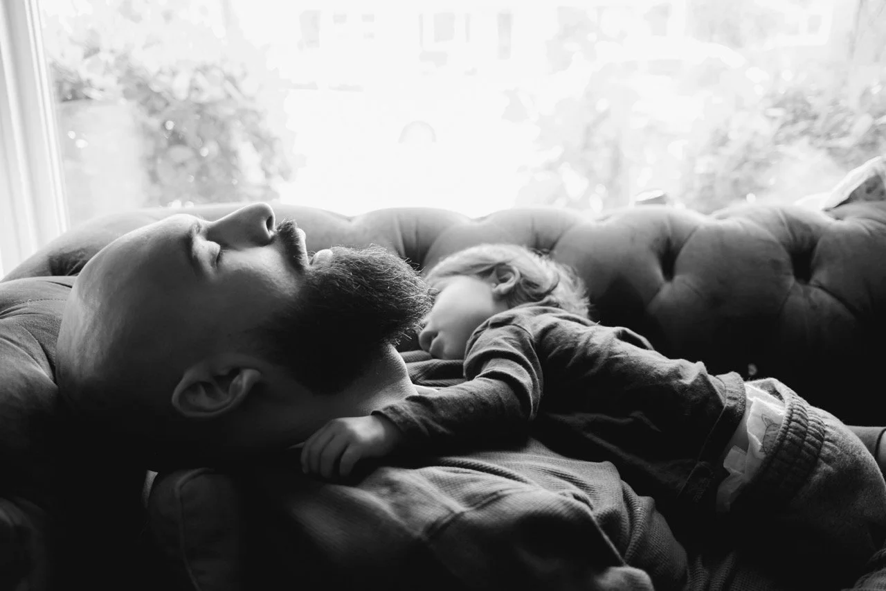 A man and a child both sleeping peacefully on a couch, with the child's head resting on the man's chest, in front of a window with natural light.