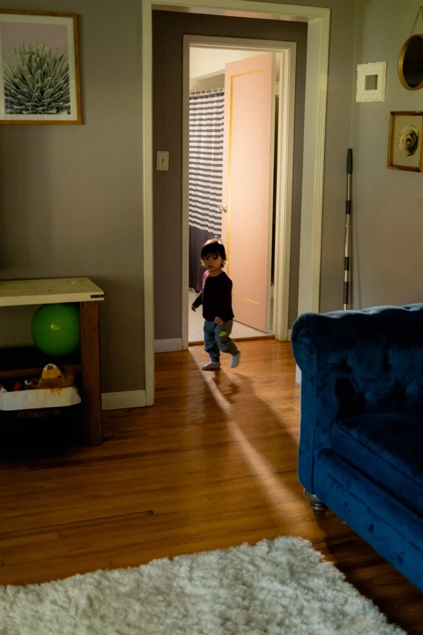 A young girl with dark hair, wearing a black jacket, jeans, and socks, walking into a room through a doorway in a cozy living room with hardwood floors and a blue sofa.