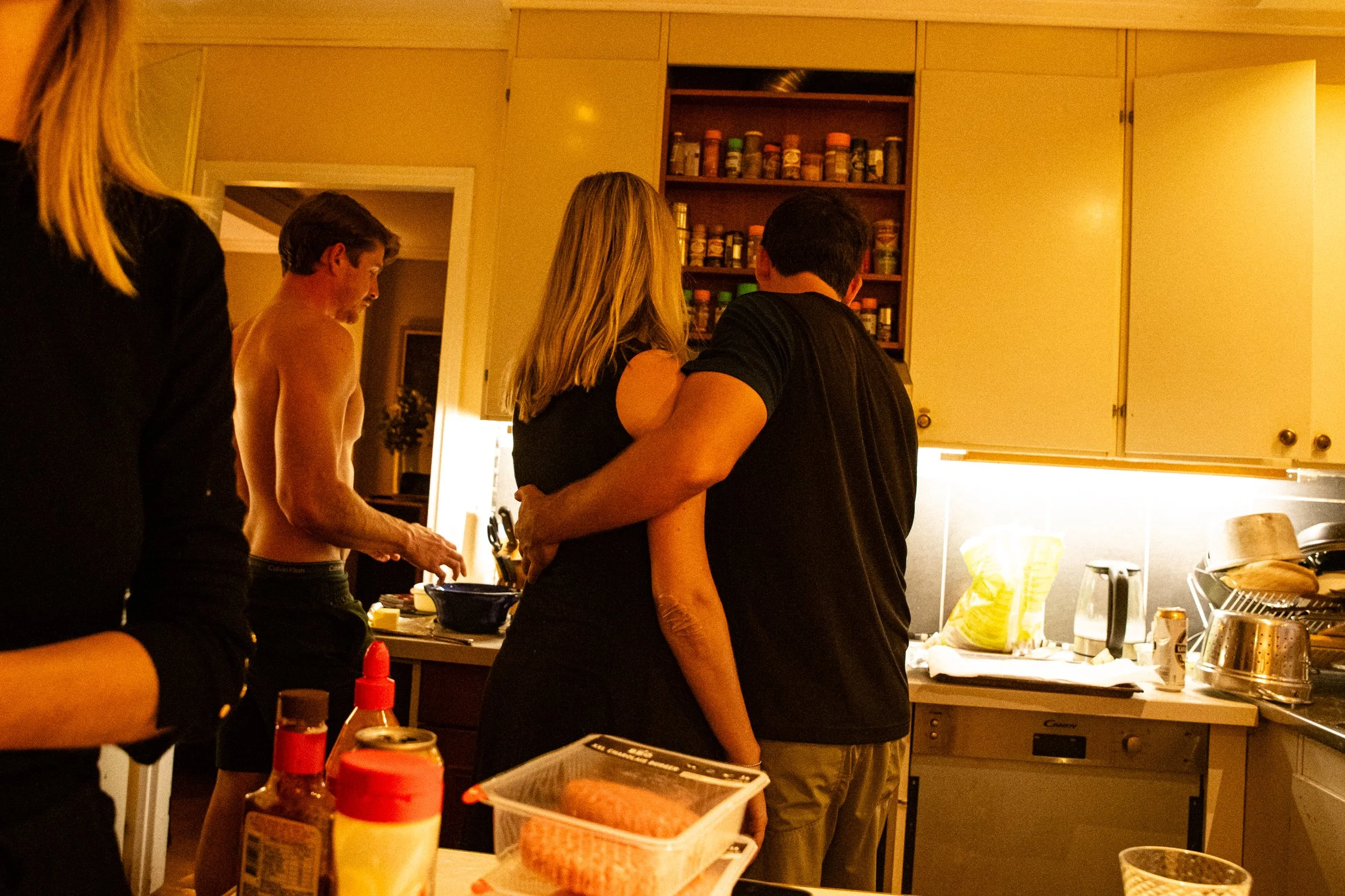 Several people are gathered around a kitchen counter, some chatting and preparing food, with a shelving unit of spices and kitchen items in the background.