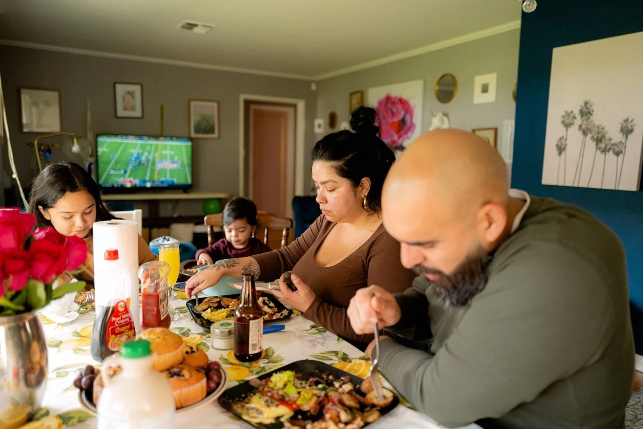 A family of four sitting at a dining table having a meal, with a TV in the background showing a sports game.
