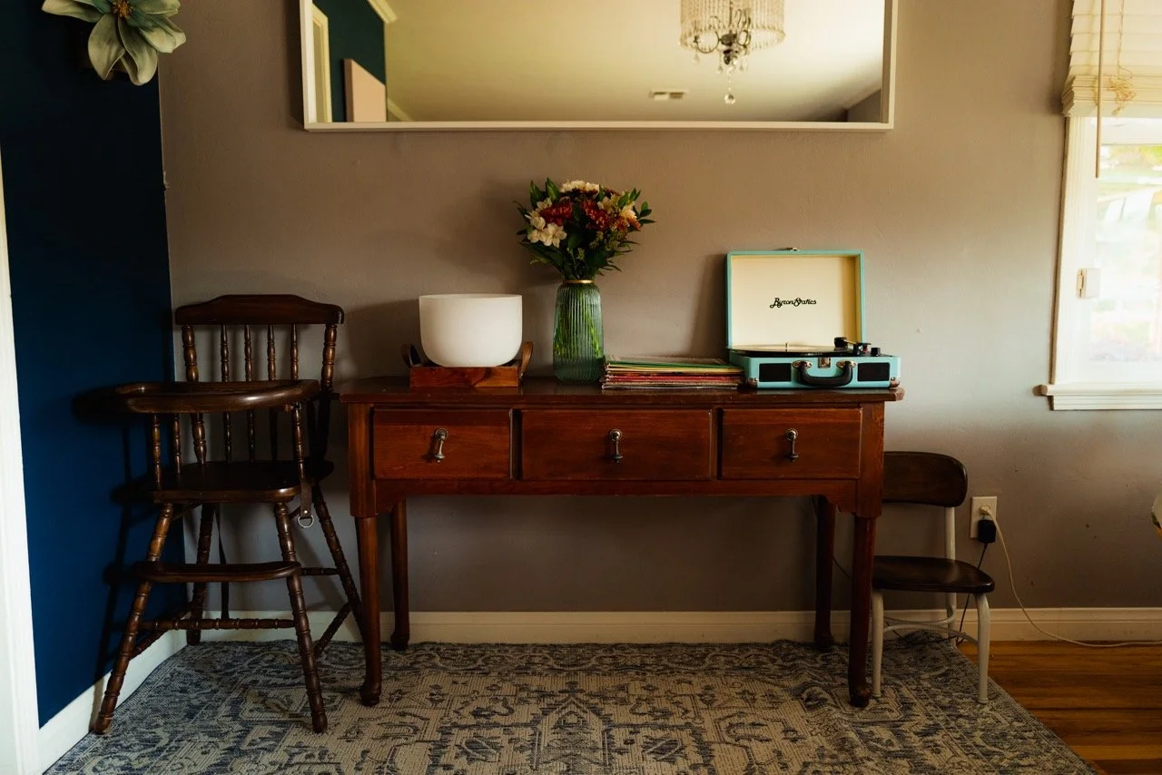 Interior of a room with a wooden console table against the wall, decorated with a green vase with flowers, a white bowl, and a portable record player. To the left of the table, there are two wooden stools stacked. A mirror hangs above the table, and 