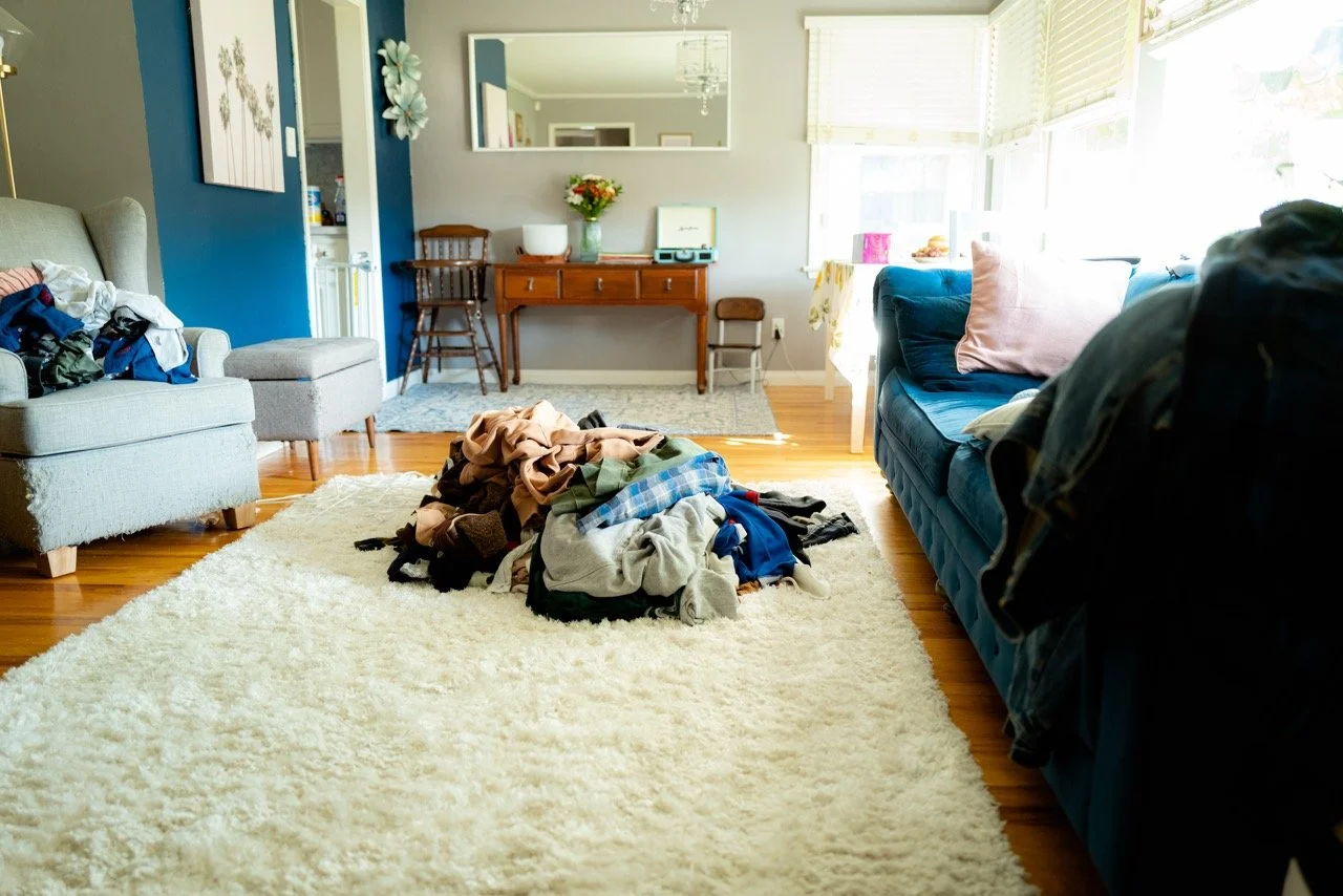 A living room with a pile of clothes on a white rug, a light gray armchair with more clothes, a blue couch with pillows, and a small table with a flower vase near the window.