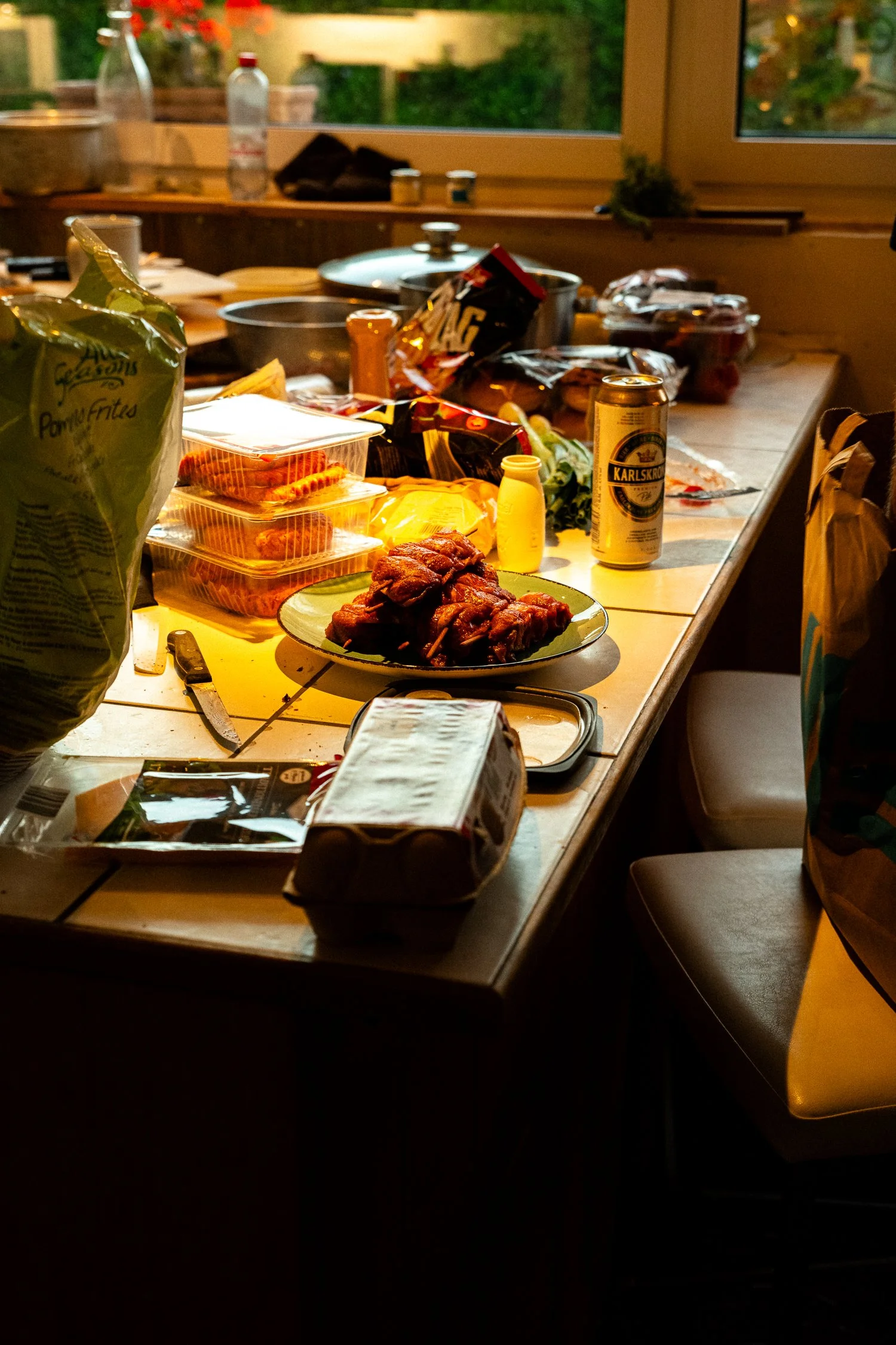 A cluttered kitchen or dining table with food items, snacks, a can of beer, and various kitchen utensils, with a window showing a garden outside.