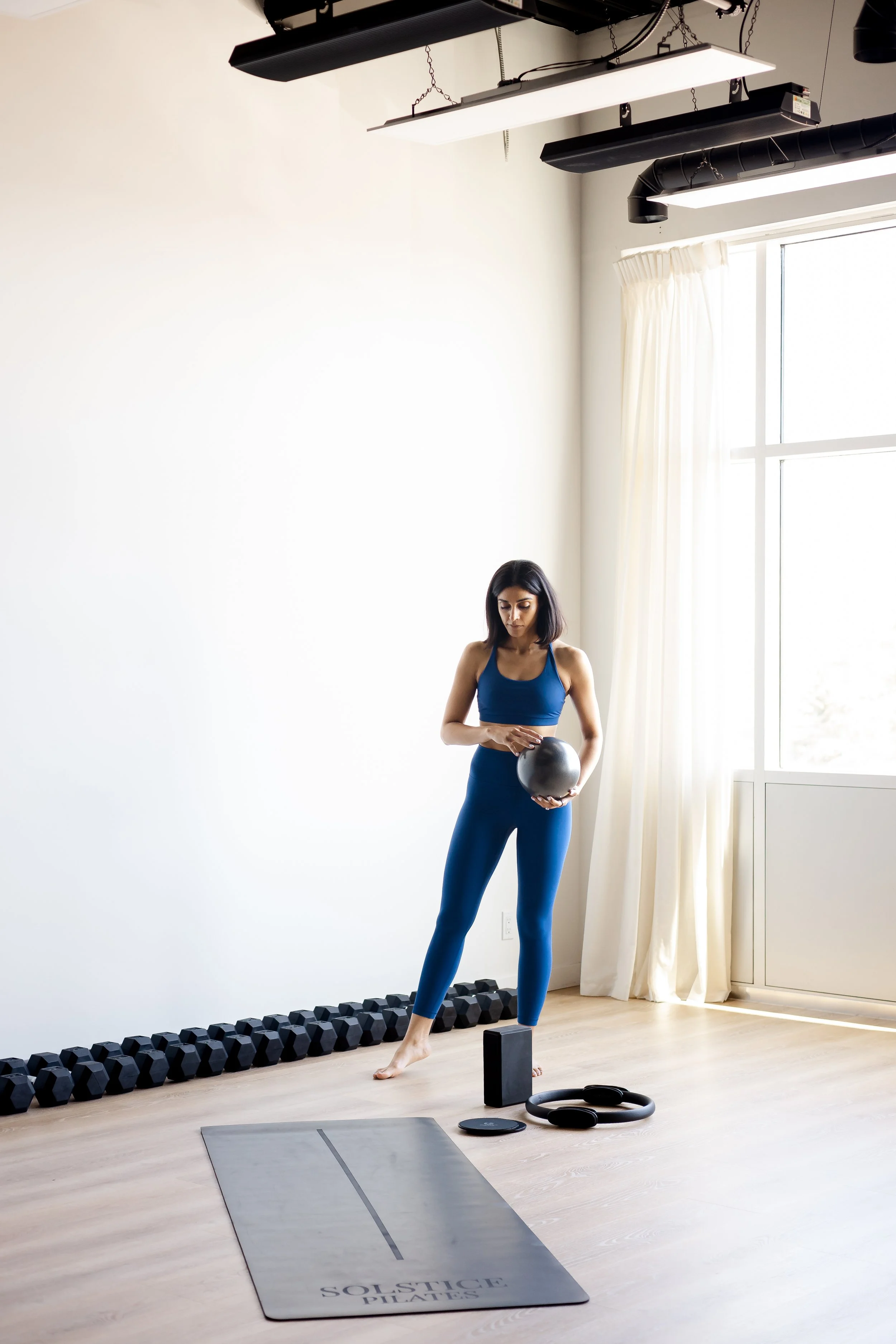 A woman in blue workout clothes holding a kettlebell in a well-lit yoga studio with large window, white walls, and fitness equipment on the floor.