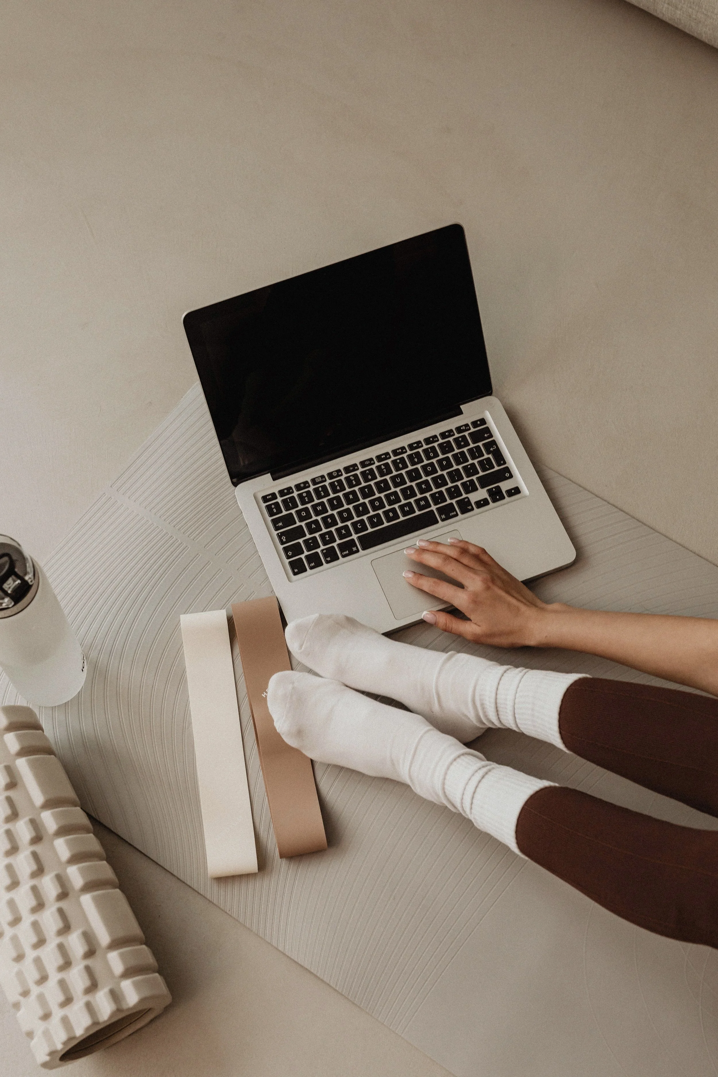 Person wearing white socks, brown pants, using a laptop on a textured beige surface, with books and a water bottle nearby.