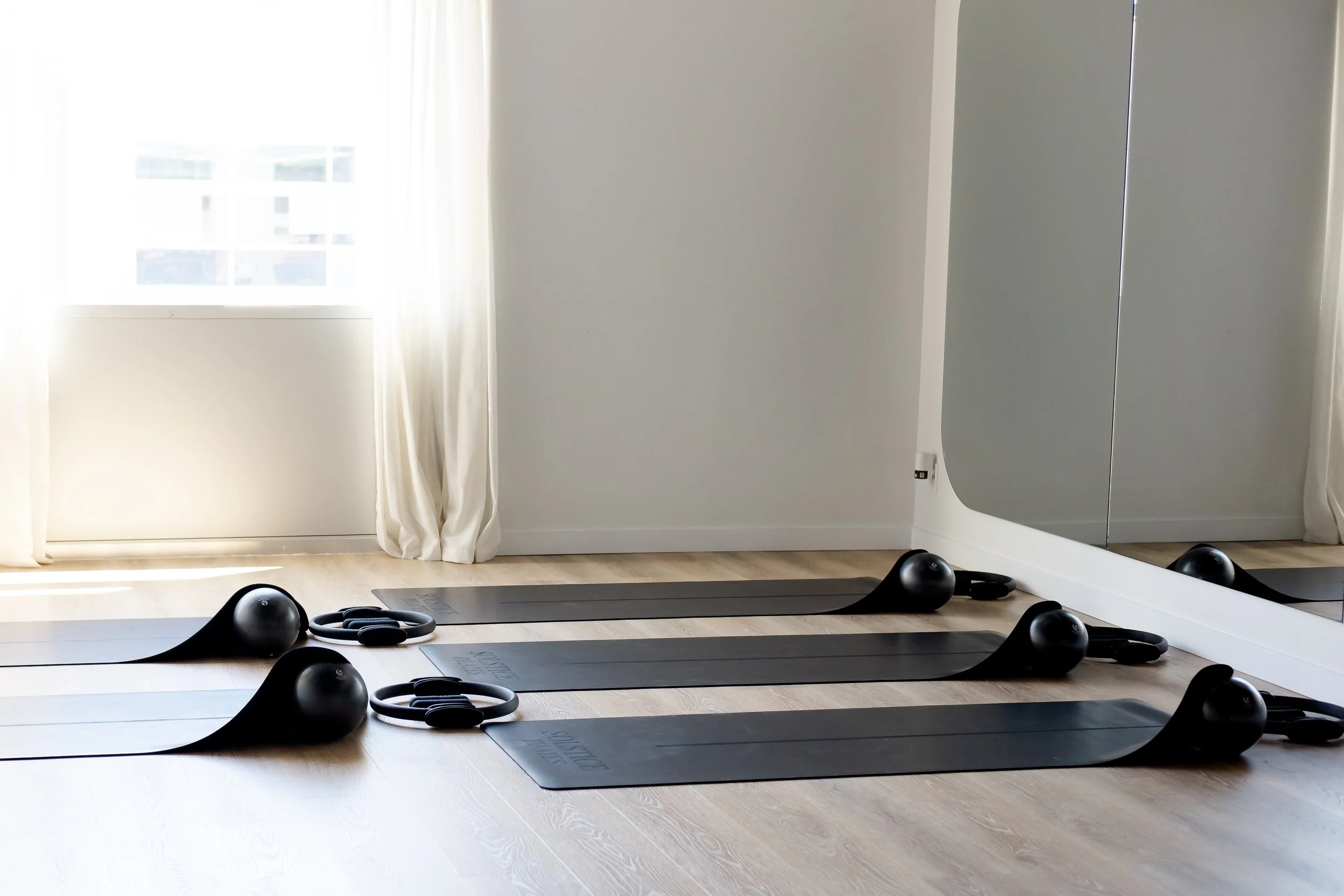 A home gym setup on a wooden floor with black exercise mats, kettlebells, and resistance bands. There is a large mirror on the right wall and a window with white curtains letting in natural light.