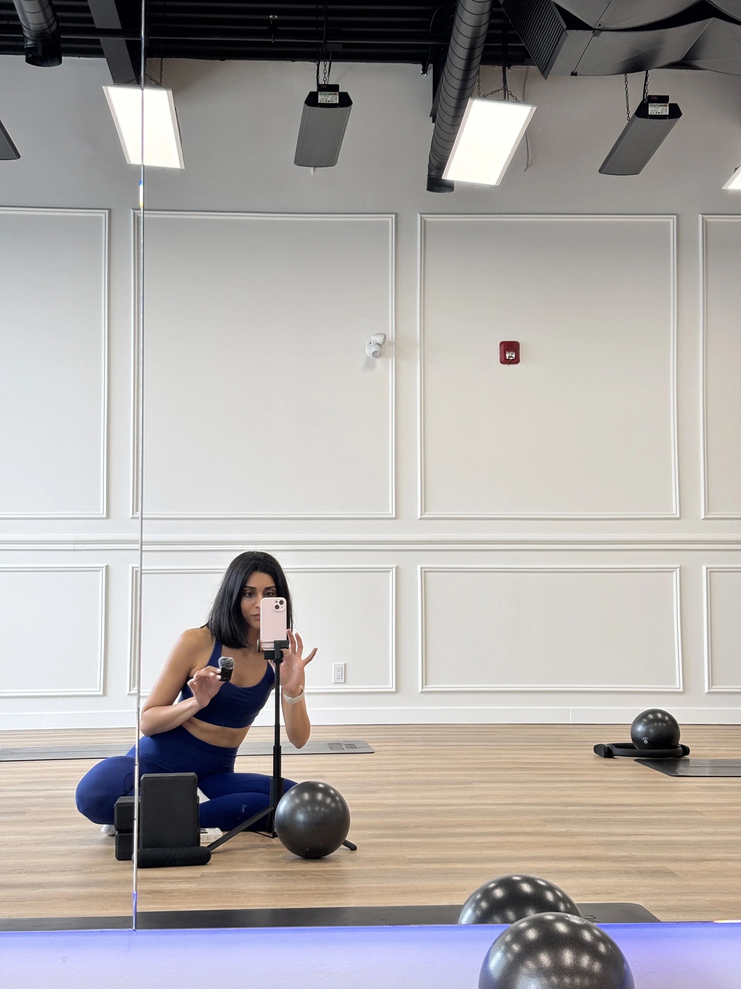 A woman with black hair in a ponytail wearing a navy sports bra and blue leggings crouching in front of a mirror in a gym, holding a dumbbell in her right hand, with workout equipment including a black medicine ball and a white wall with decorative molding in the background.