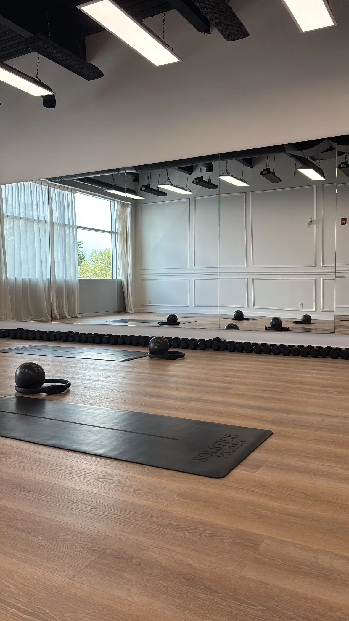 Empty fitness or yoga studio with wooden floors, black exercise mats, kettlebells, resistance bands, and a large mirror reflecting windows with curtains, bright natural light, and ceiling lights.