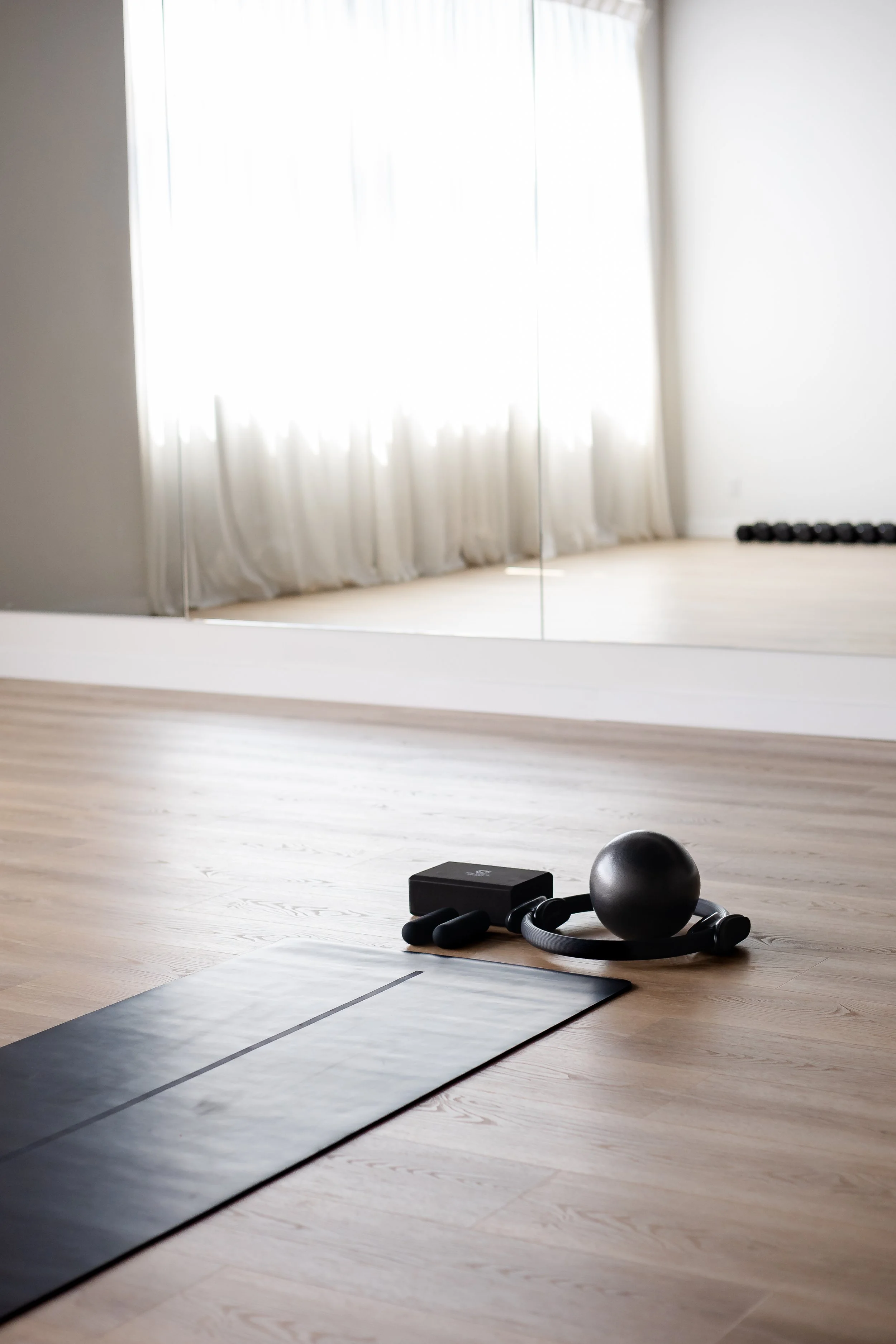A home gym with a yoga mat, a black medicine ball, a kettlebell, and gym accessories on a wooden floor near a mirror. Bright natural light through curtains illuminates the space.
