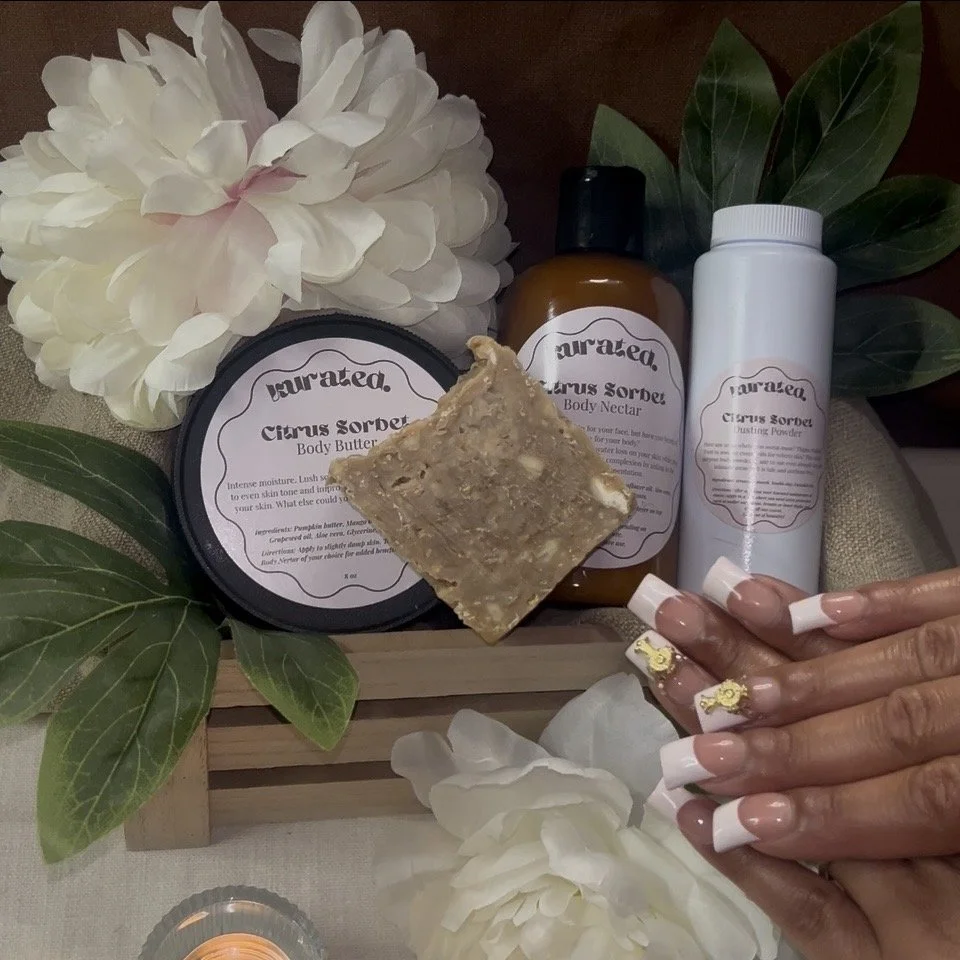 Collection of skincare and body care products with white hydrangea flowers and green leaves, including a container of body butter, a bottle of body nectar, and a bottle of body dusting powder, placed on a wooden tray, with a person's hand displaying 