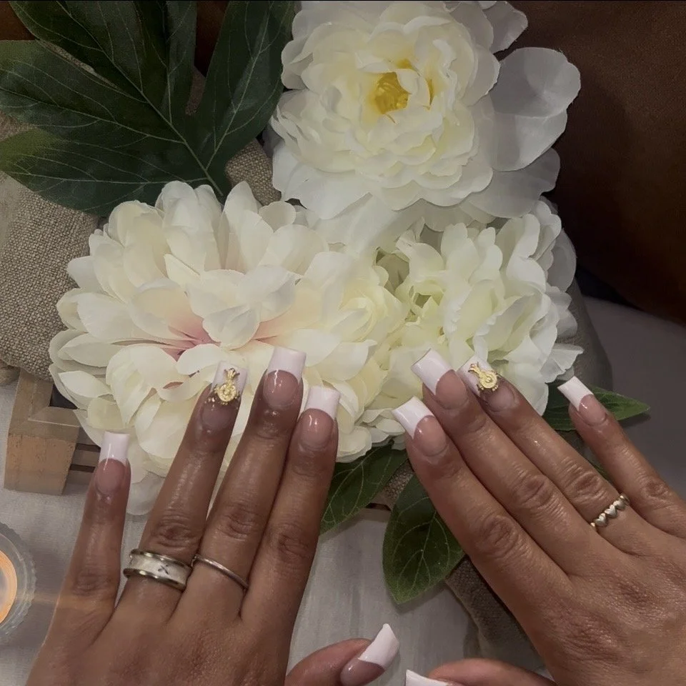 Hands with manicured nails and rings, placed on a table with white and cream flowers, green leaves, and a lit candle.