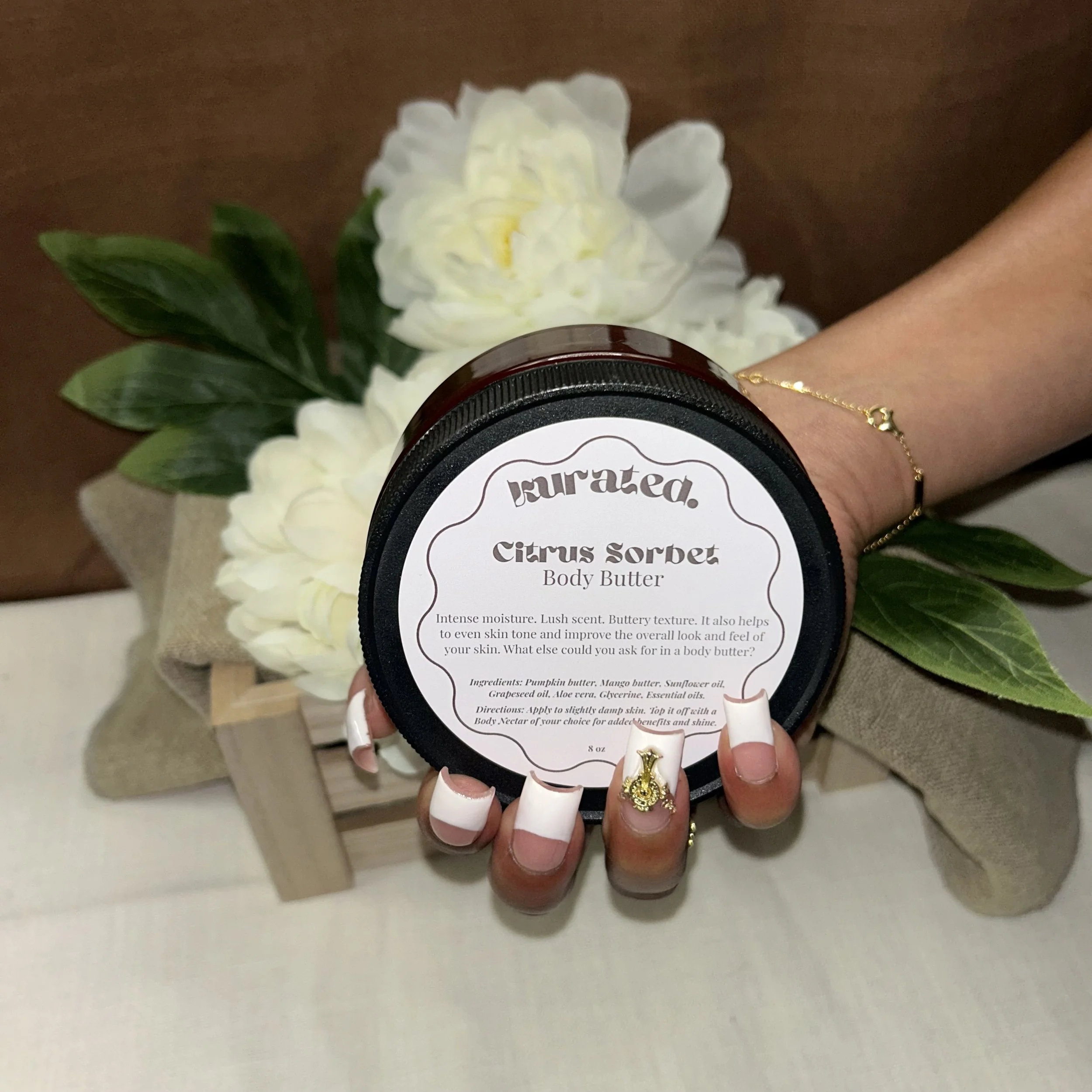 Close-up of a hand with manicured nails holding a container of body butter labeled 'Citrus Sorbet' with a background of white flowers and green leaves.
