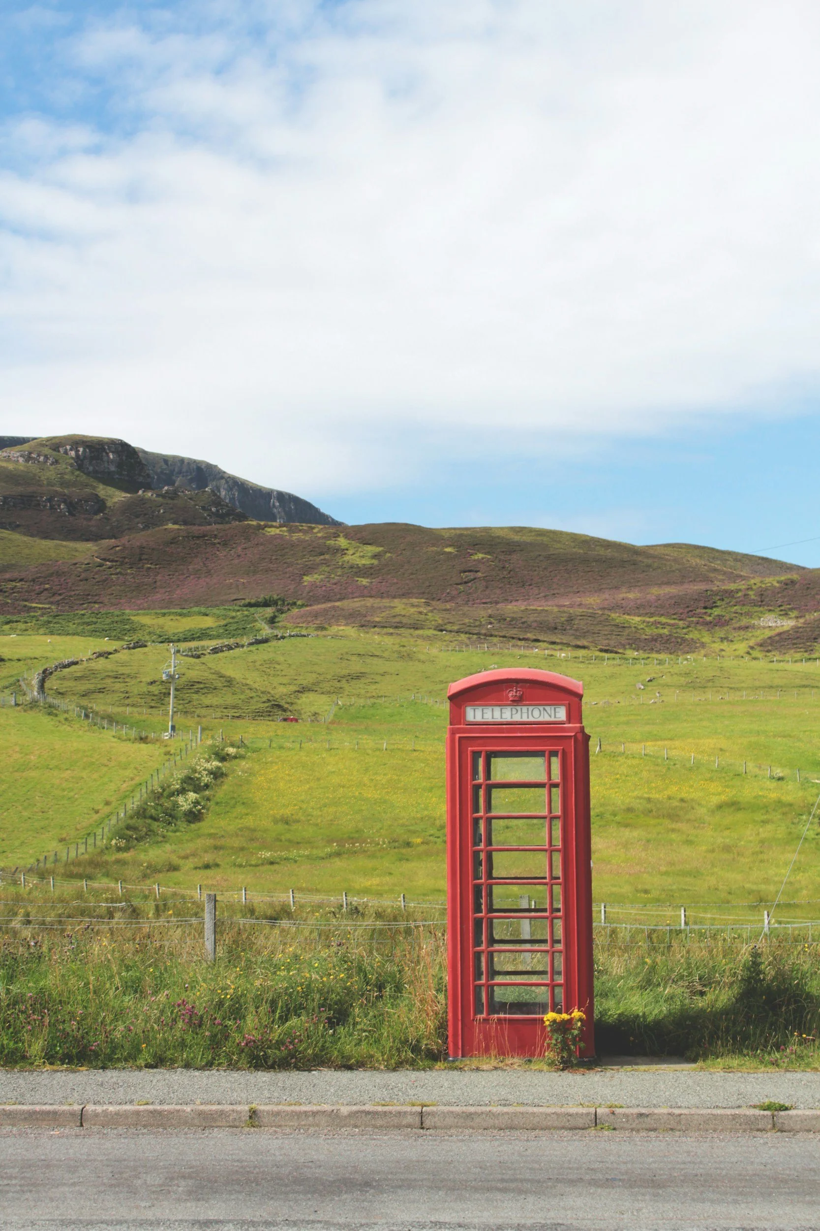 A red telephone booth on a roadside with green fields, hills, and a partly cloudy blue sky in the background. UK England weather chatgpt uk mot check royal mail tracking airbnb berko Berkhamstead Art art local artist original drawing painting