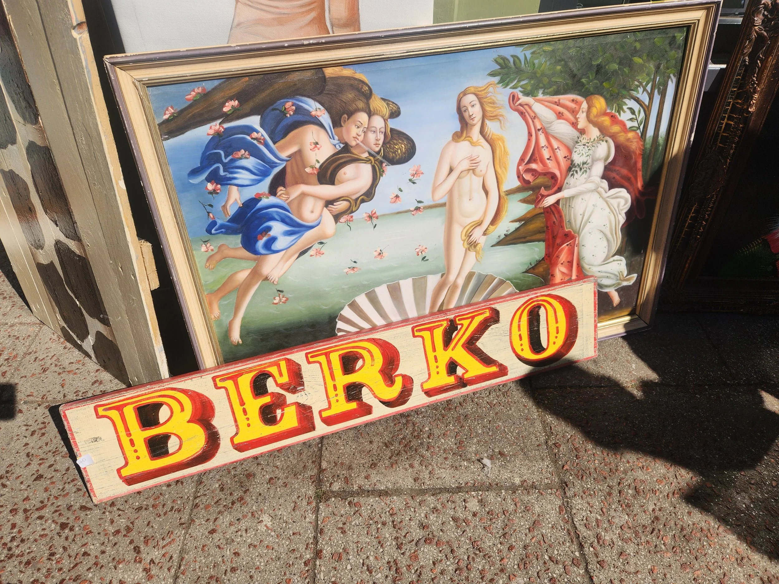 A framed renaissance-style painting of three women, one nude, standing in a shell under a tree with pink flowers, and a sign with yellow and red text "BERKO". Sandro Bottichelli botticelli The birth of Venus Berko Berkhamstead Art Fair mermaids