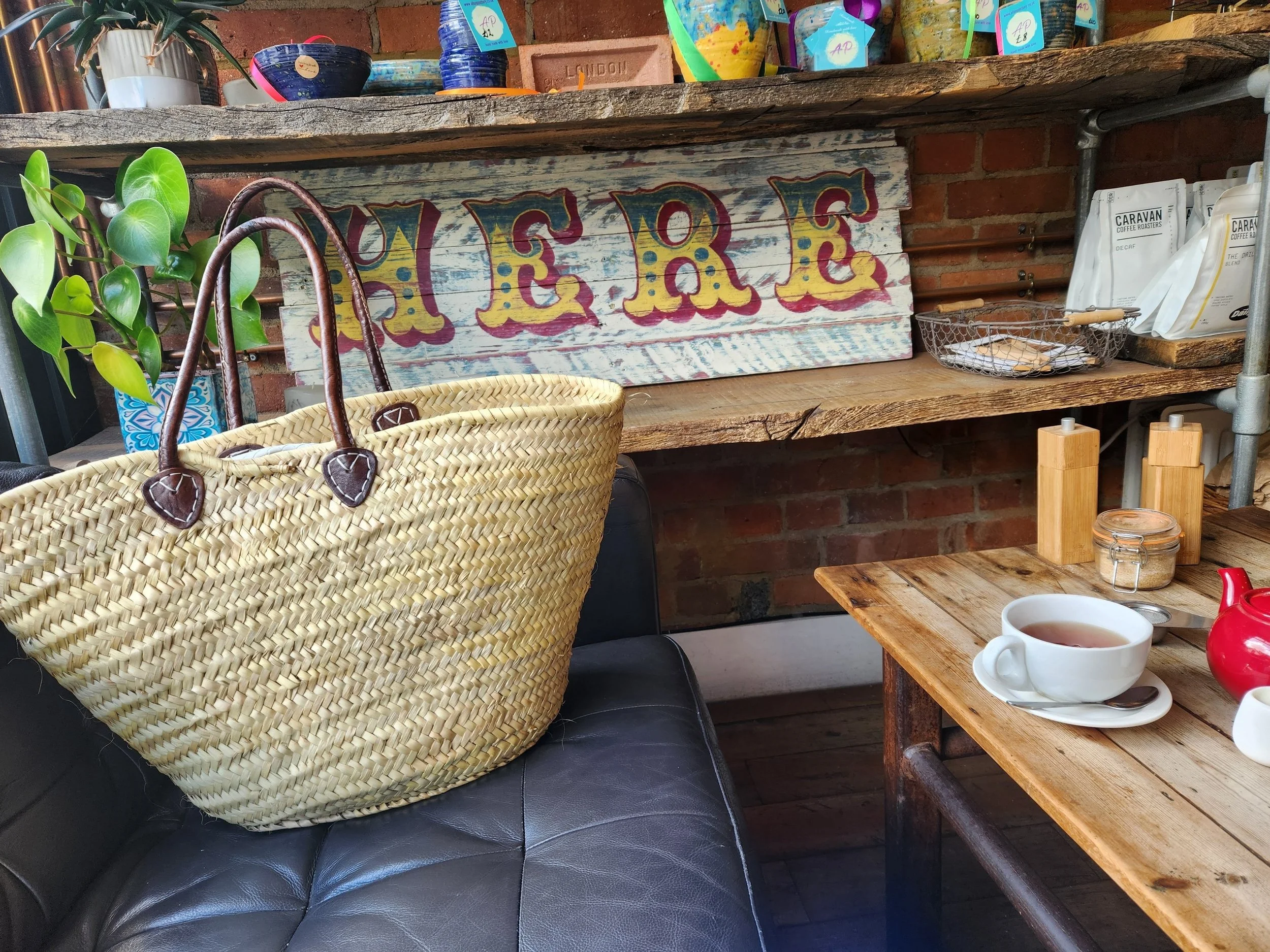 Cozy café setting with a black leather couch, a basket bag on it, a wooden table with a cup of tea, a shelf with colorful pots, a large painted wooden sign reading 'HERE', potted plants. Berko Berkhamstead Mermaid Brunch restaurants Art England