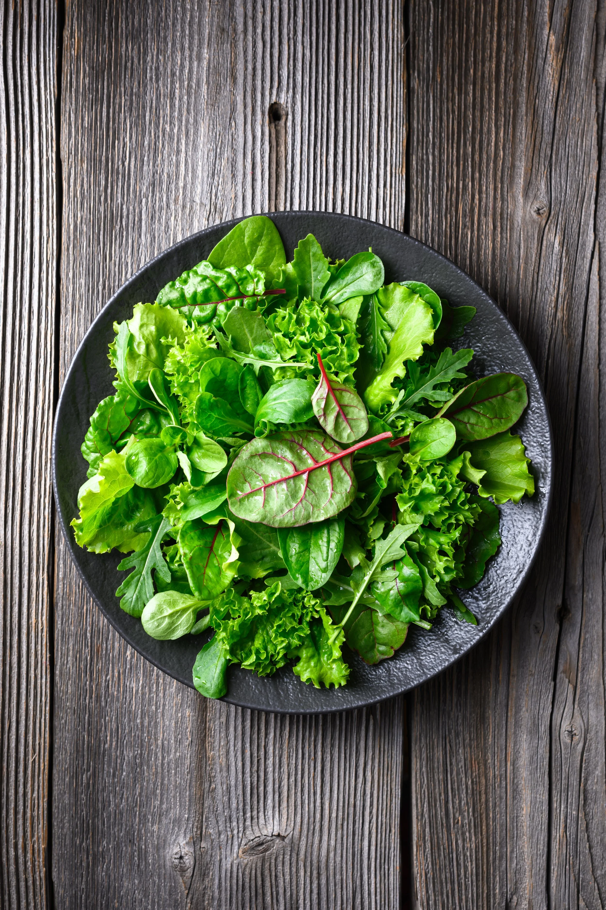 A black bowl filled with assorted fresh green salad greens on a rustic wooden table.