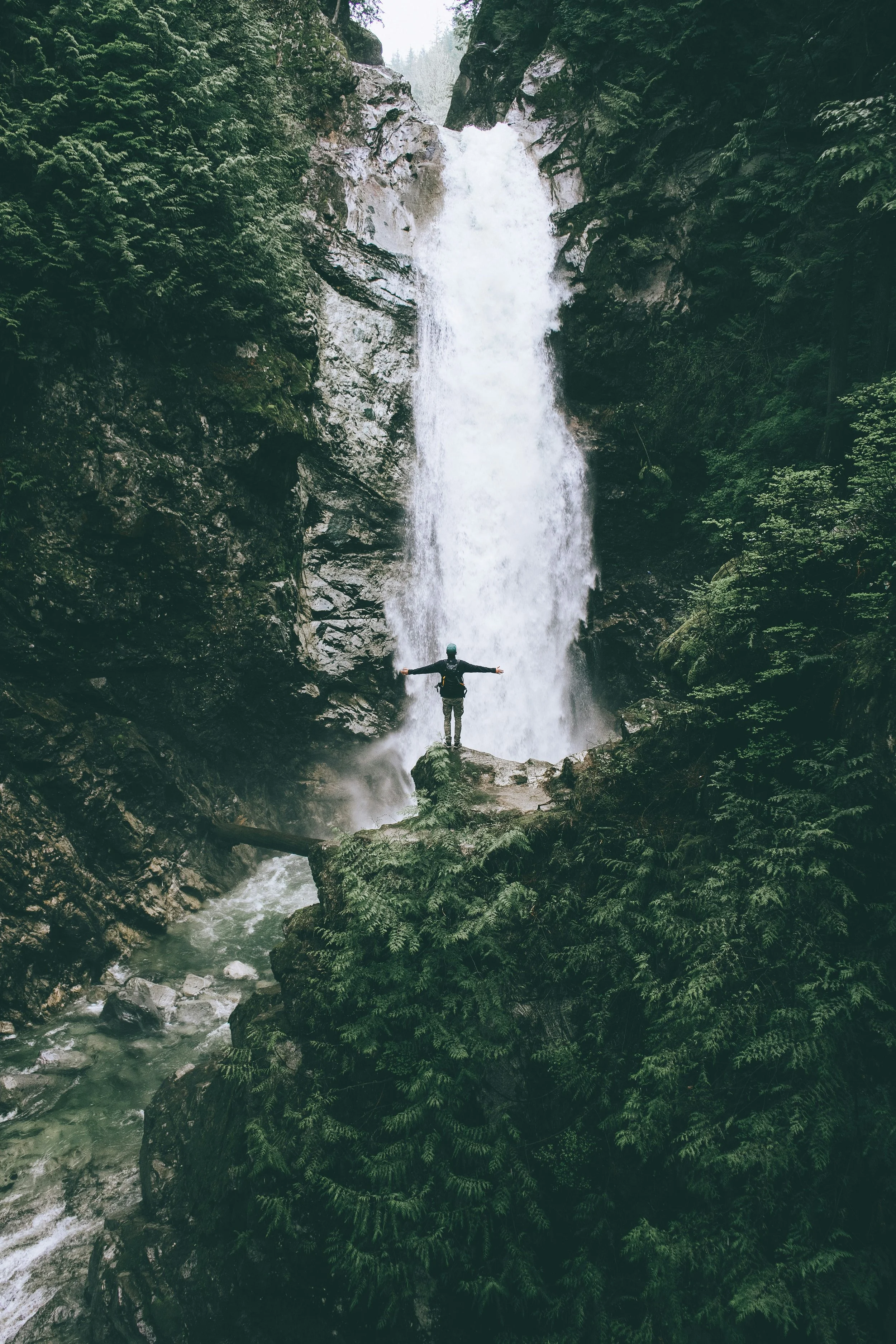 Person standing on rocks with arms outstretched near a tall waterfall in a lush, green forest.