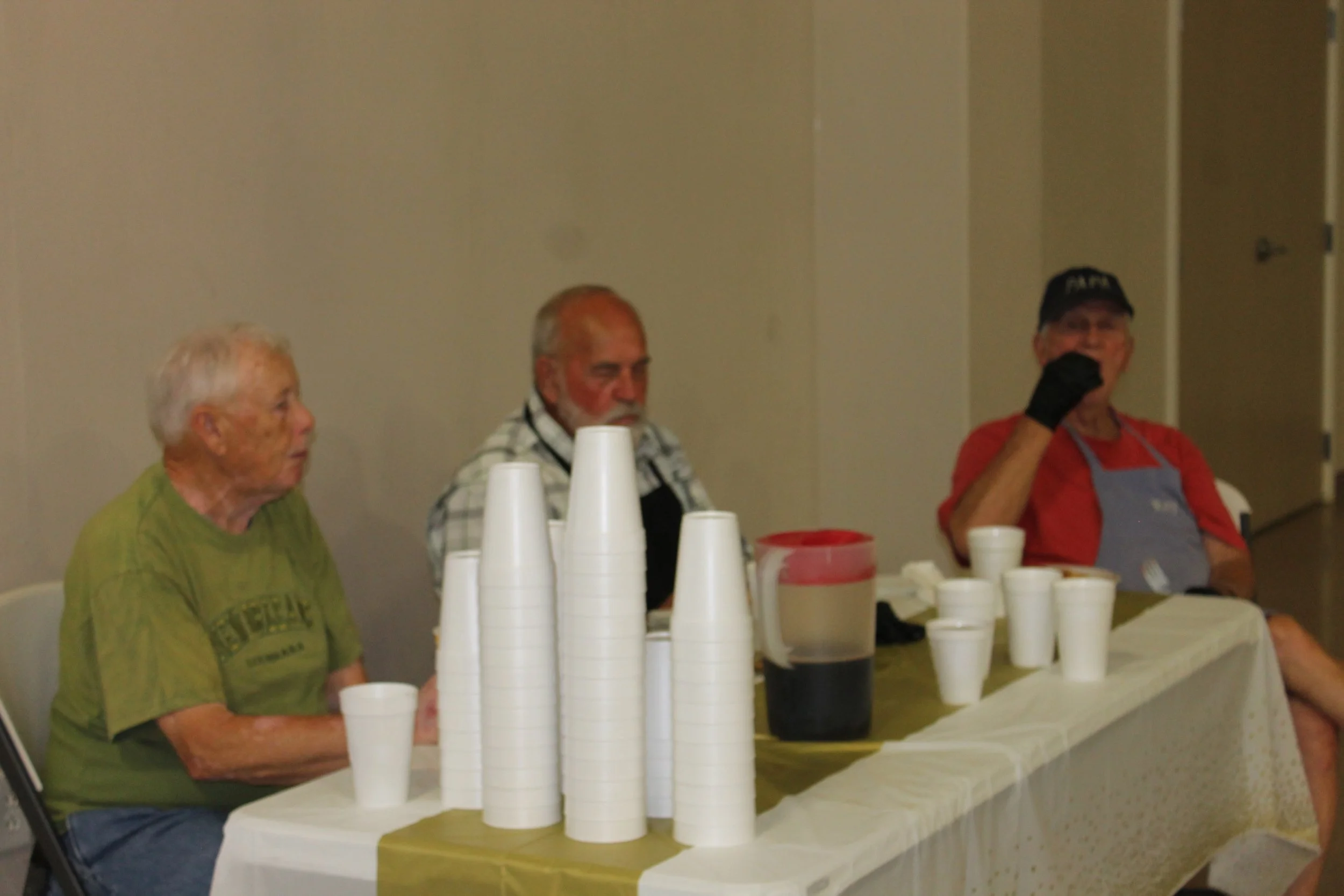 Three elderly men are sitting at a table with stacked white cups and pitchers. One man is wearing a green T-shirt, the second is wearing a plaid shirt, and the third is wearing a red shirt and a black cap. The room has plain beige walls and a closed door.