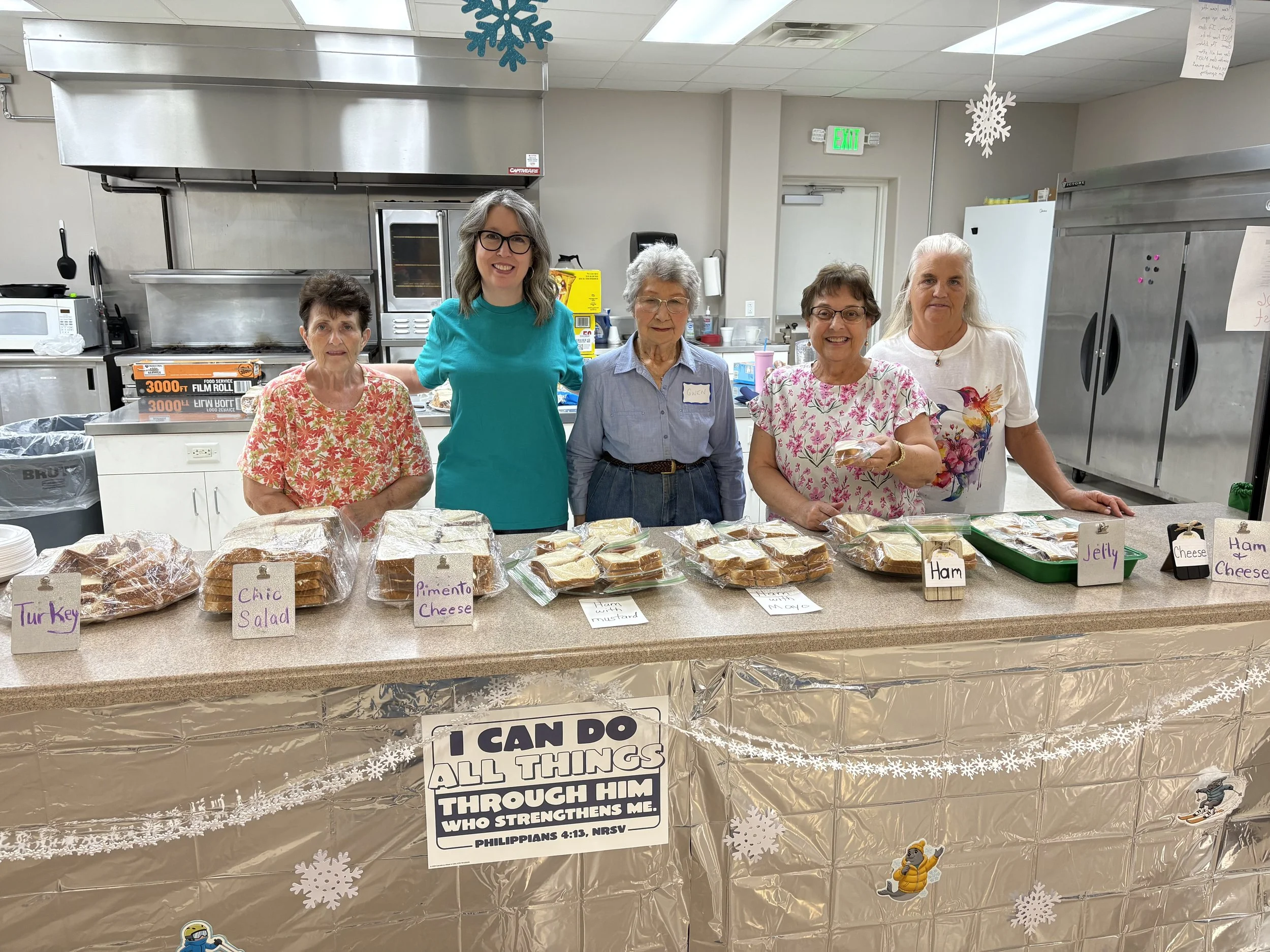 A group of five women standing behind a table with various foods, such as turkey, chips, pimento cheese, ham with mustard, jelly, cheese, and ham cheese, labeled with handwritten signs. The women are smiling in a kitchen with a stainless steel oven, refrigerator, microwave, and snowflake decorations hanging from the ceiling.