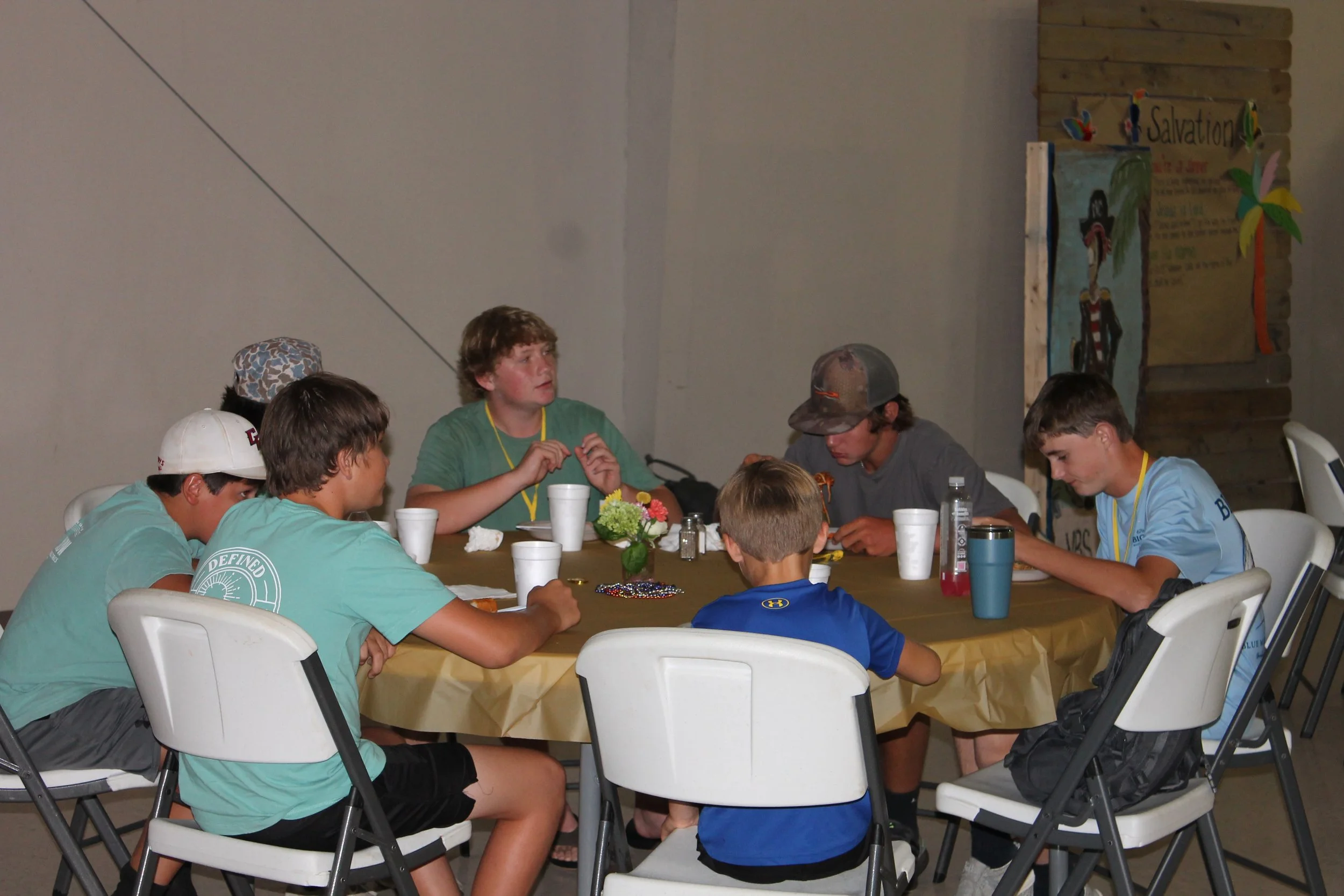 A group of young people sitting around a table at a gathering or meeting, with some wearing colorful shirts and a backdrop that includes a decoration with a sign that says 'Salvation' and artwork of a person.