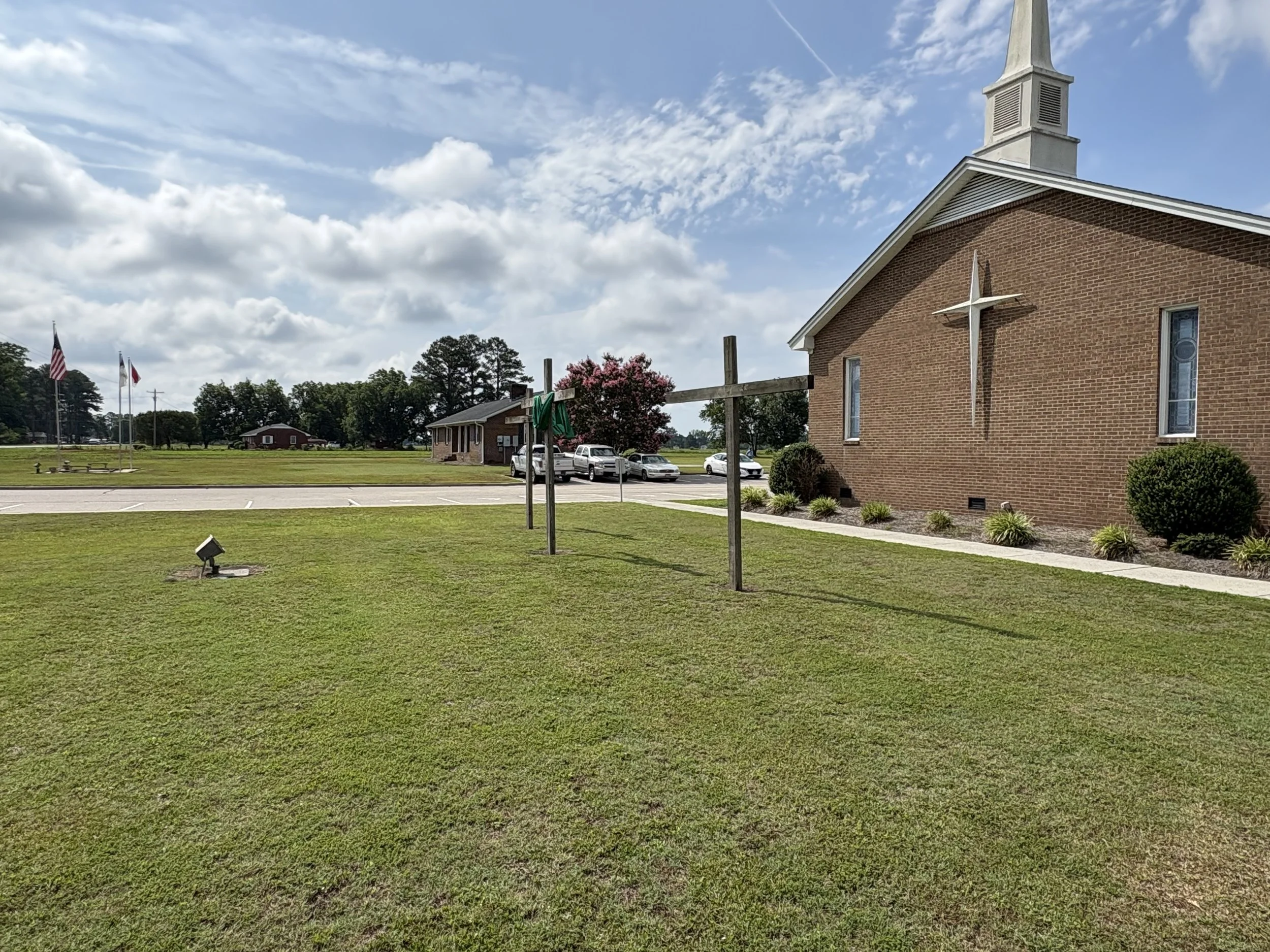A church building with a steeple and a large cross on the brick wall. In front, there are flagpoles with American and other flags, a small parking lot, and a grassy area with a few plants and a floodlight. The sky is partly cloudy.