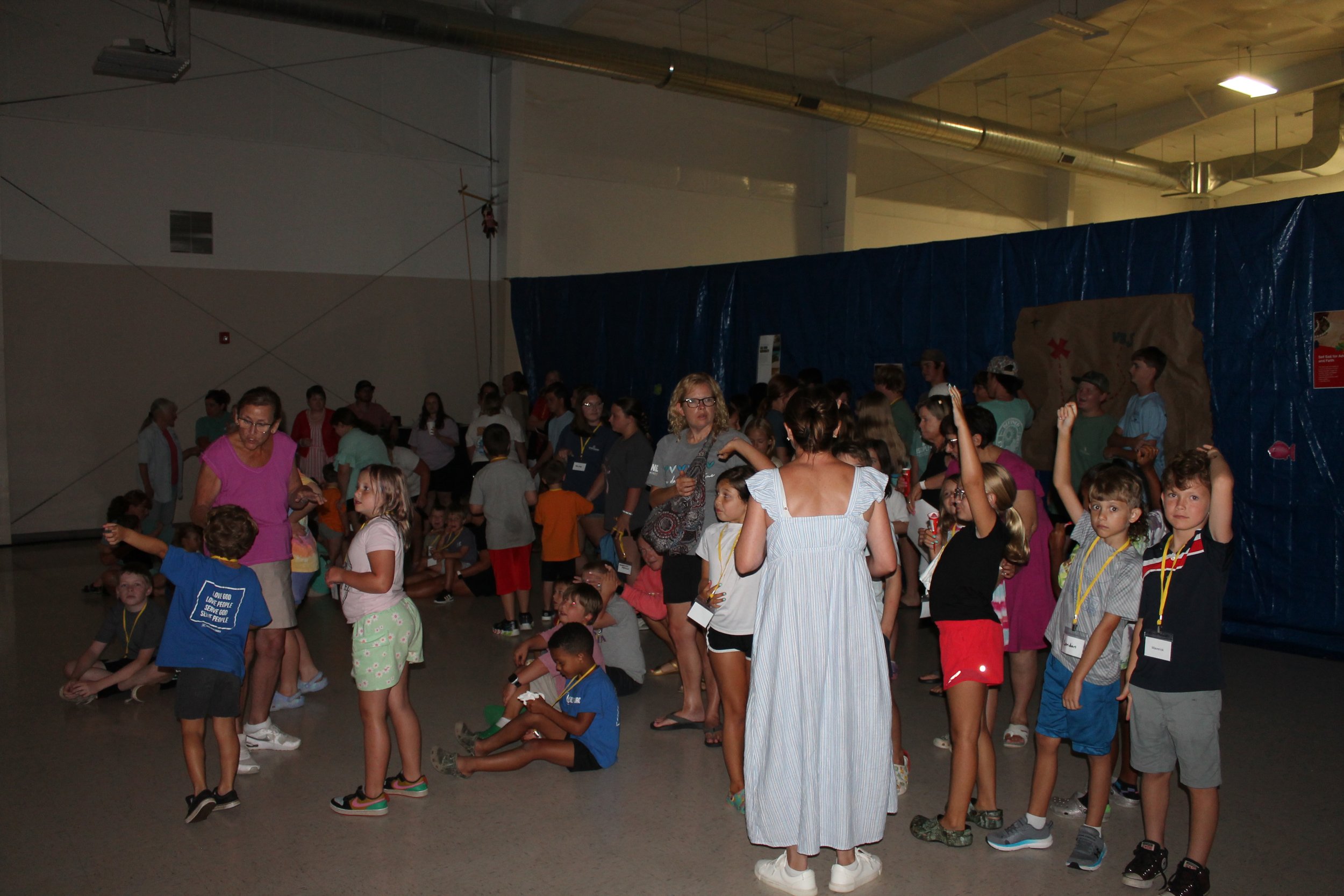 A large group of children and adults gathered indoors, likely at a recreational or educational event, with some children sitting and others standing, engaged in various activities or conversations.