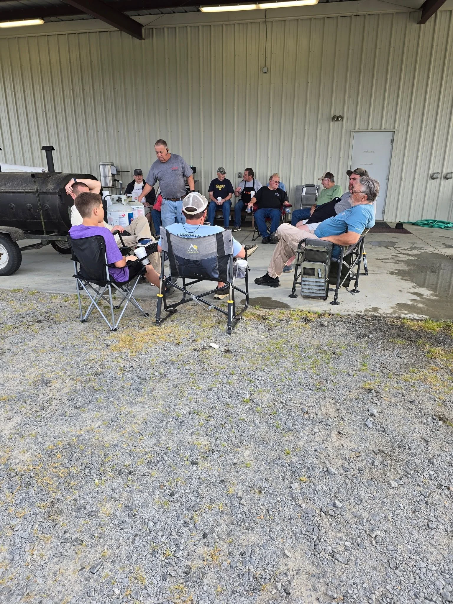 Group of people sitting on chairs and a bench outside a large metal building, some eating, others talking, with a grill nearby.