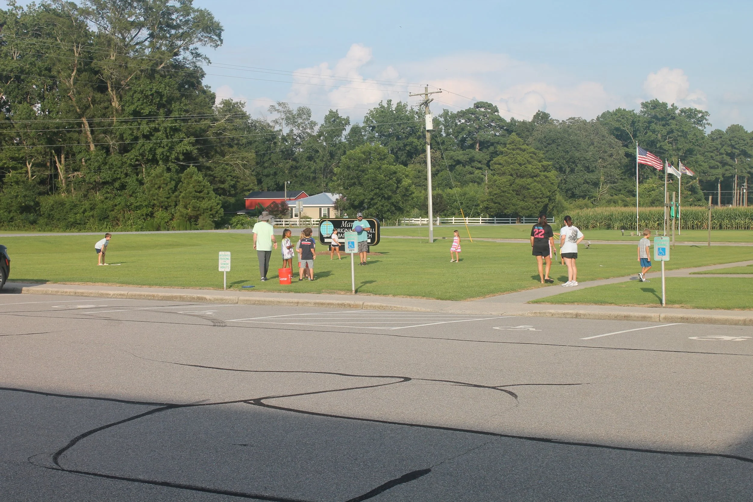 A parking lot with visible parking spaces and in the background, a grassy area with several people, including children, participating in outdoor activities. There are flags flying on flagpoles, including the American flag, and a sign indicating a disabled parking zone. The scene includes trees, a small building, and utility poles, under a partly cloudy sky.