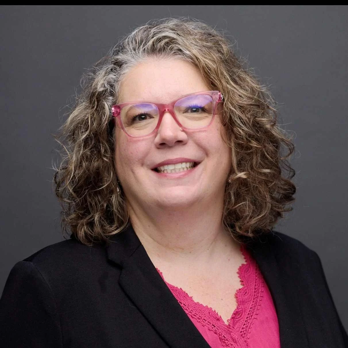 A woman with curly, shoulder-length hair, wearing pink glasses, a black blazer, and a pink blouse, smiling against a dark gray background.