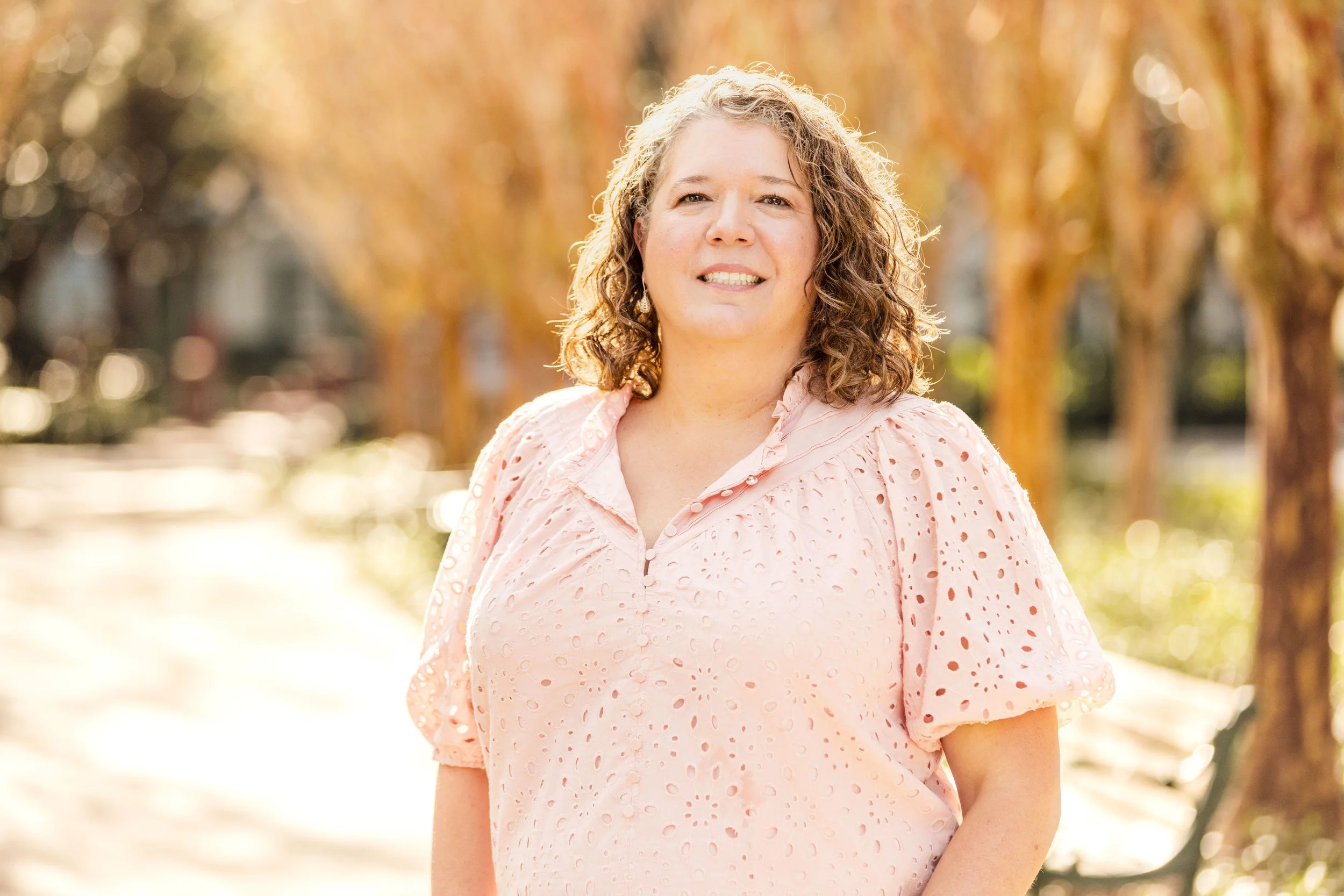 A woman with curly hair smiling outdoors during autumn, wearing a light pink blouse with eyelet details.