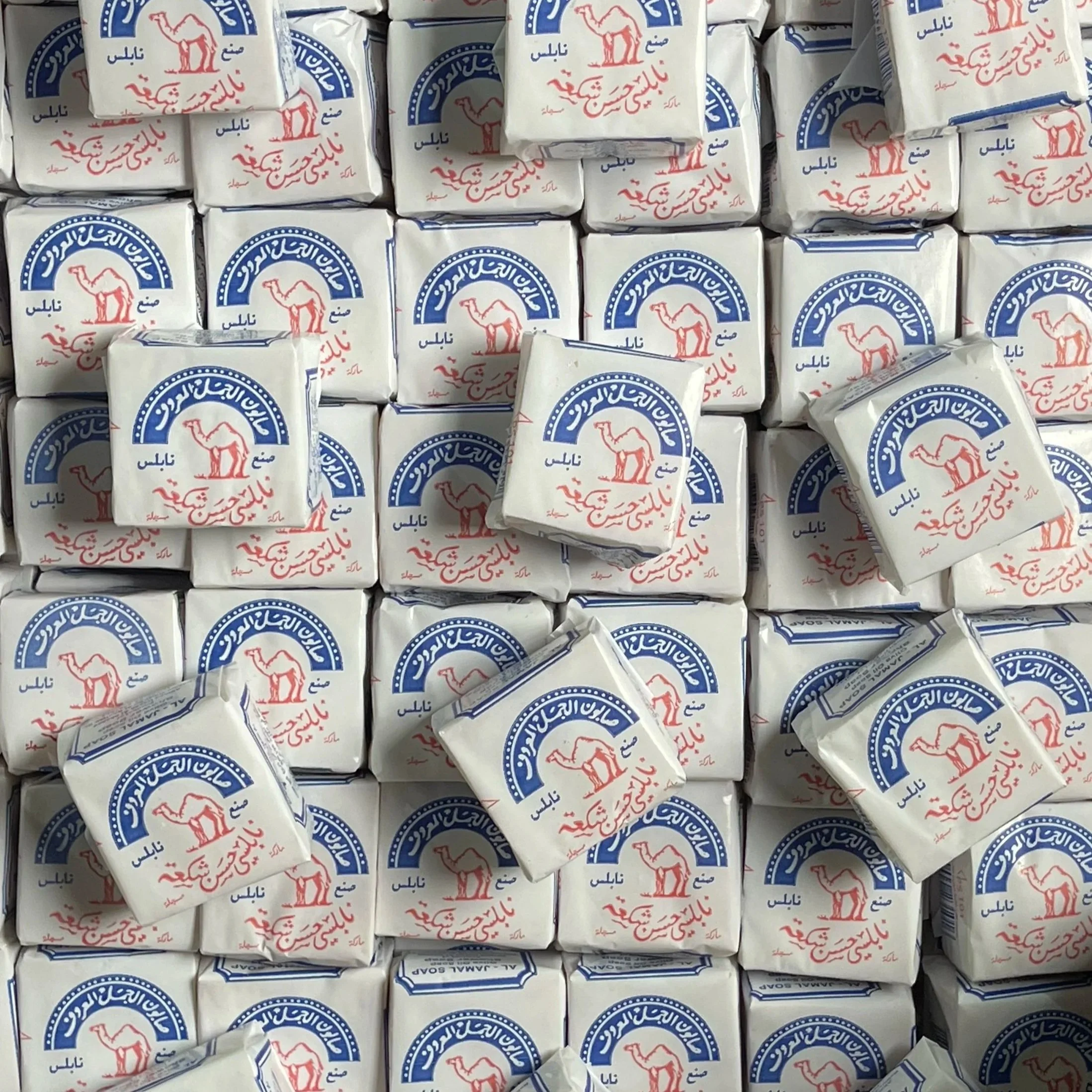 A close-up of multiple bars of soap wrapped in white packaging with red and blue Arabic text and a logo featuring a camel.
Packaging of Al-Jamal Nabulsi Olive Oil Soap.