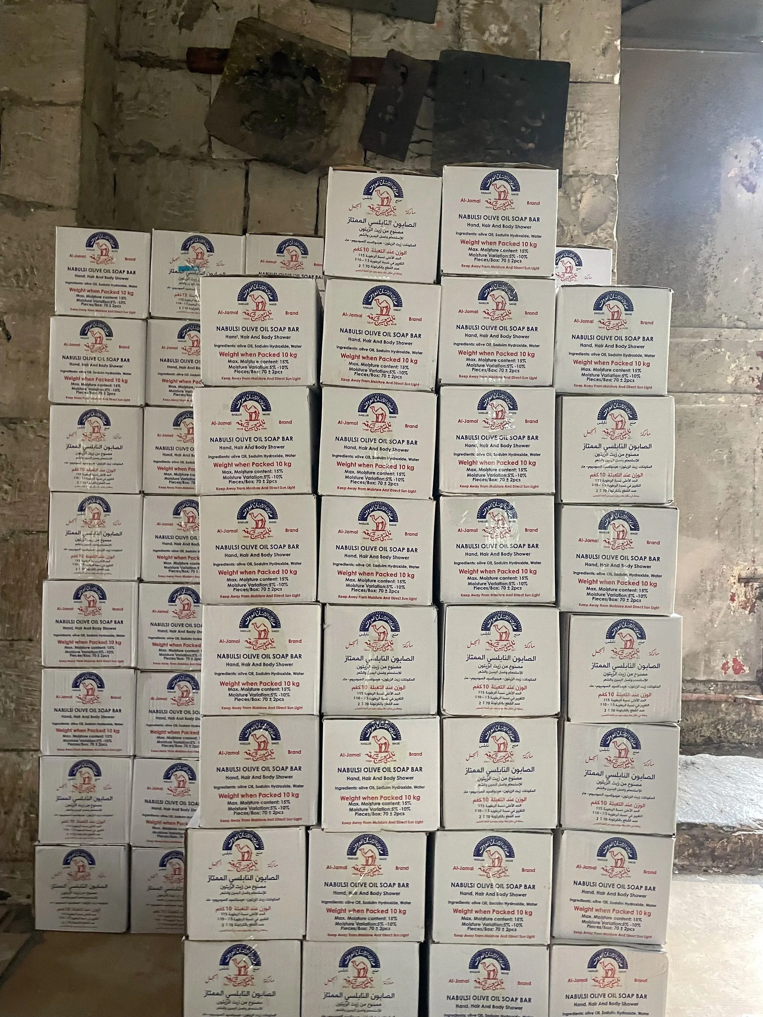 Multiple white boxes of Al-Jamal Nabulsi Olive Oil Soap Bar stacked against a rough stone wall.