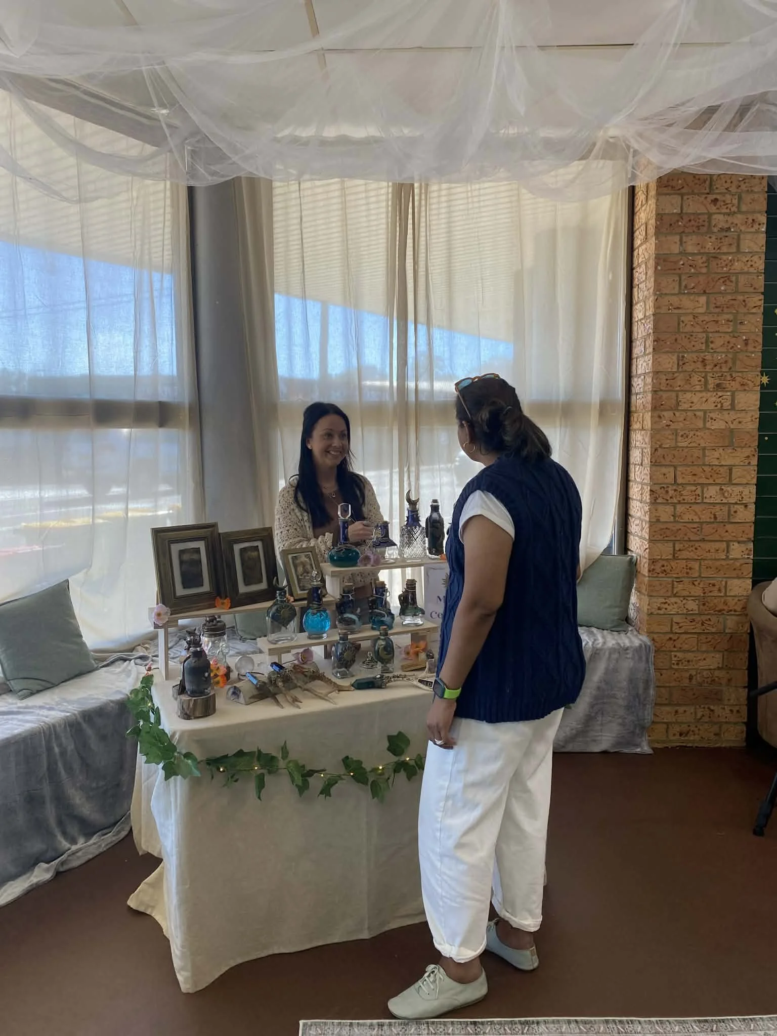 A woman in white pants and a navy blue vest is standing in front of a display of decorative bottles, picture frames, and jewelry at a craft fair or market. She is talking to a smiling woman behind the table, which is decorated with pictures, flowers, and greenery, under a sheer canopy with flowing fabric. The setting is inside a room with a brick wall and large windows covered with curtains.