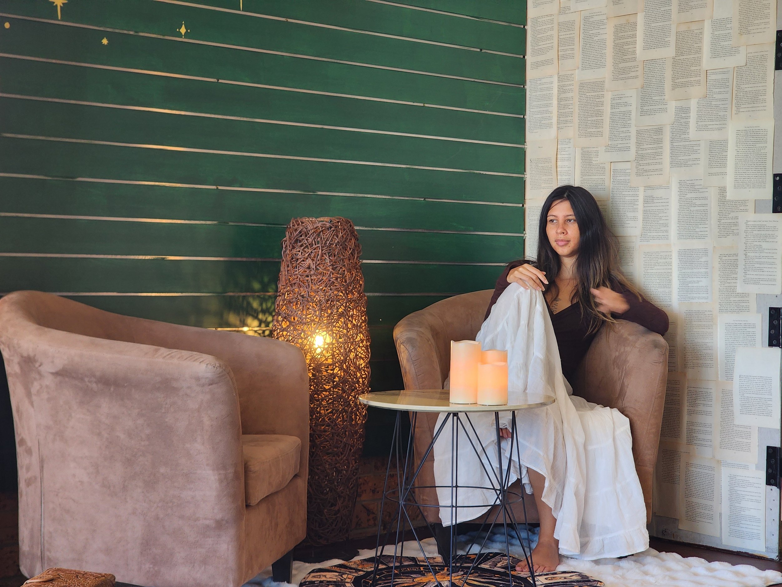 A woman sitting on a beige armchair next to a small round table with lit candles and a decorative wicker lamp inside a cozy room with green panels on one wall and pages of text on another wall.