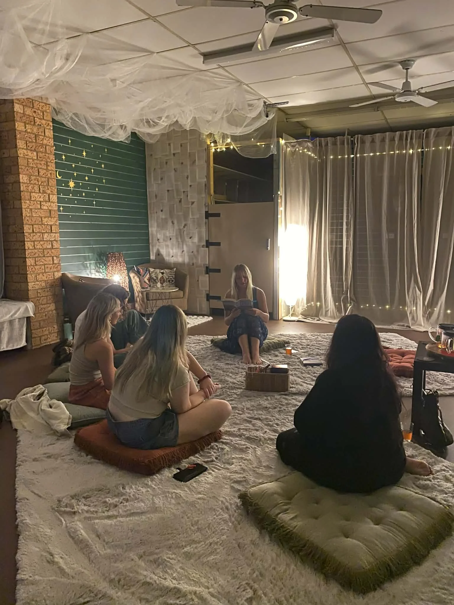 Group of people sitting on a rug in a cozy room with string lights, curtains, and decorative wall backdrops, while one woman reads aloud from a book.