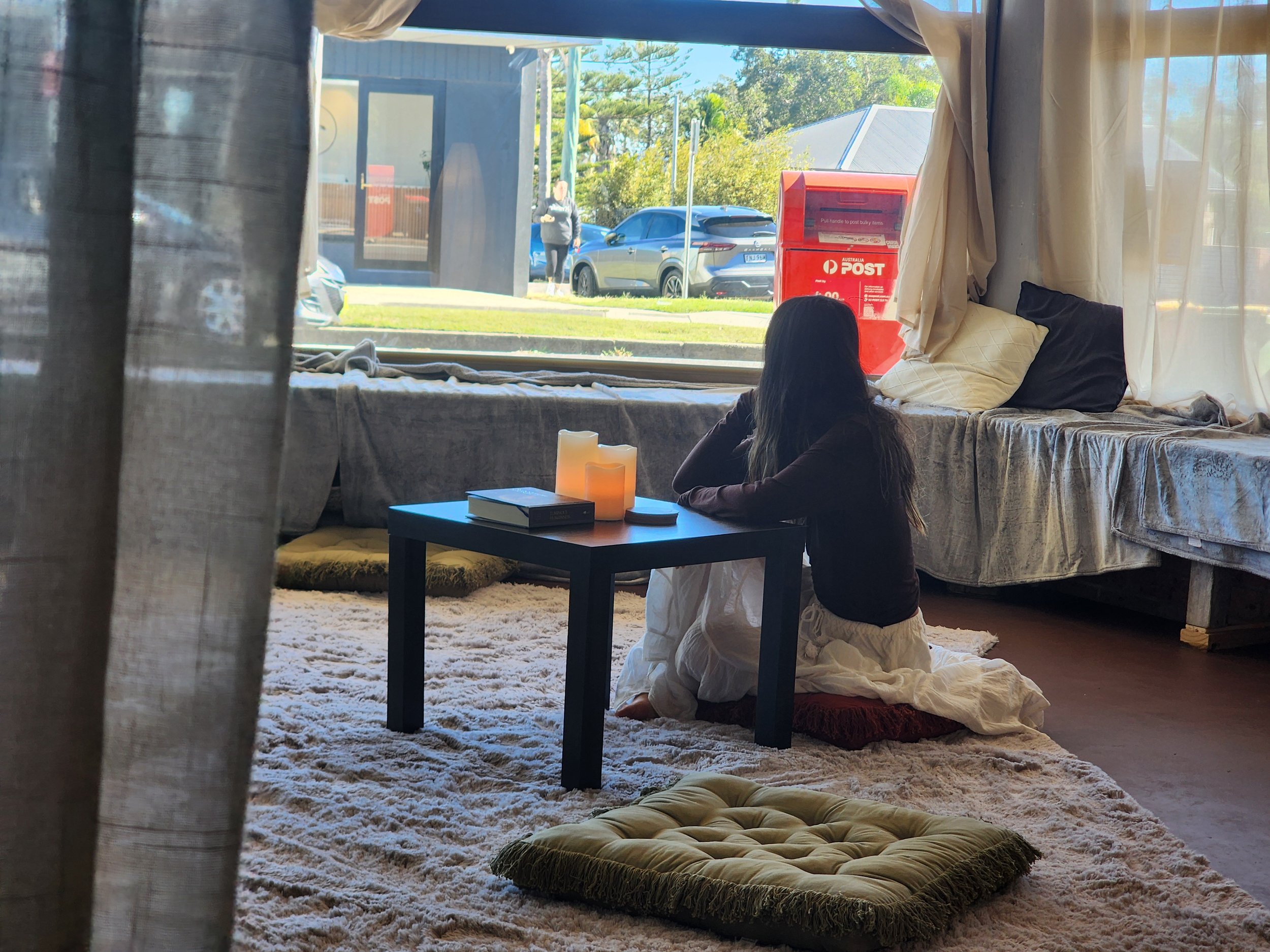 Woman kneeling in prayer on a beige cushion inside a cozy room, with sunlight streaming through the windows. Outside view shows parked cars, a person walking, trees, and a red Australia Post mailbox.