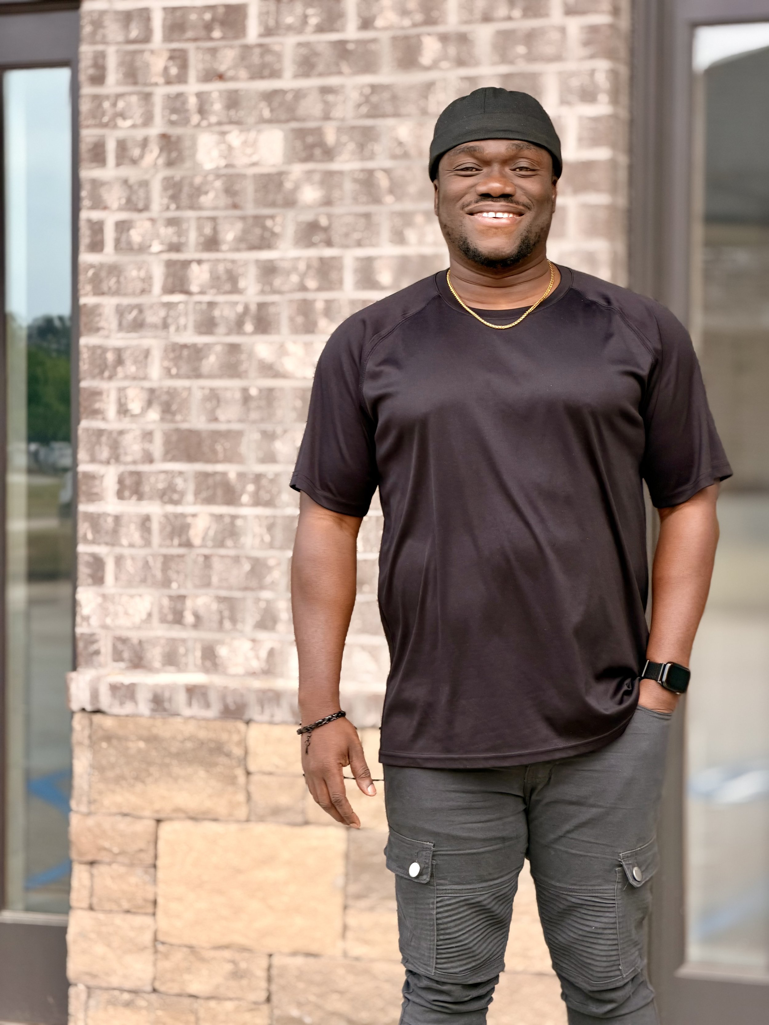 A man wearing a black cap, black t-shirt, and gray cargo pants stands outdoors near a brick wall, smiling and looking at the camera.