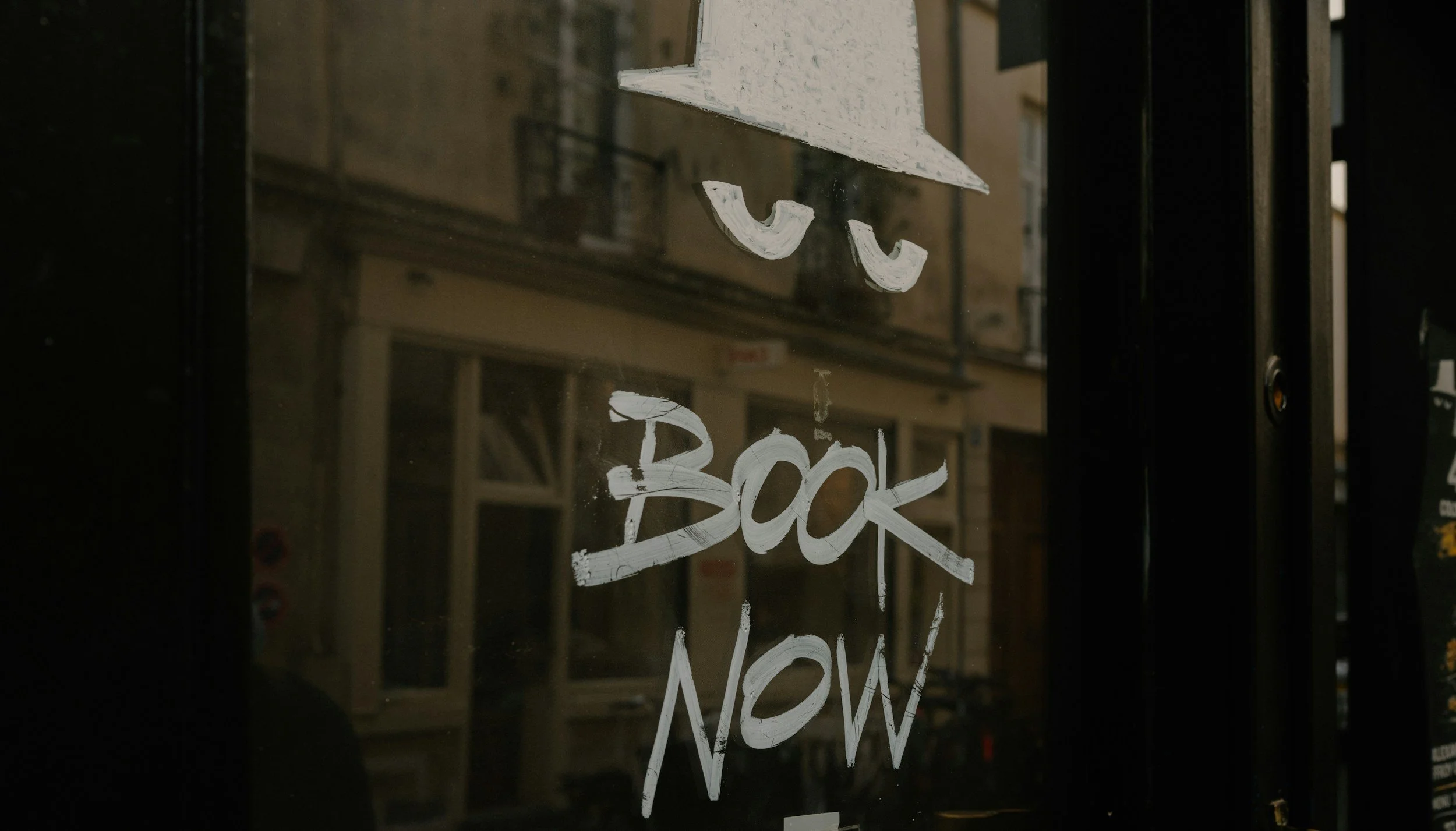 A storefront window with handwritten white text that says "We Suck Now". The reflection of buildings and a street can be seen in the glass.