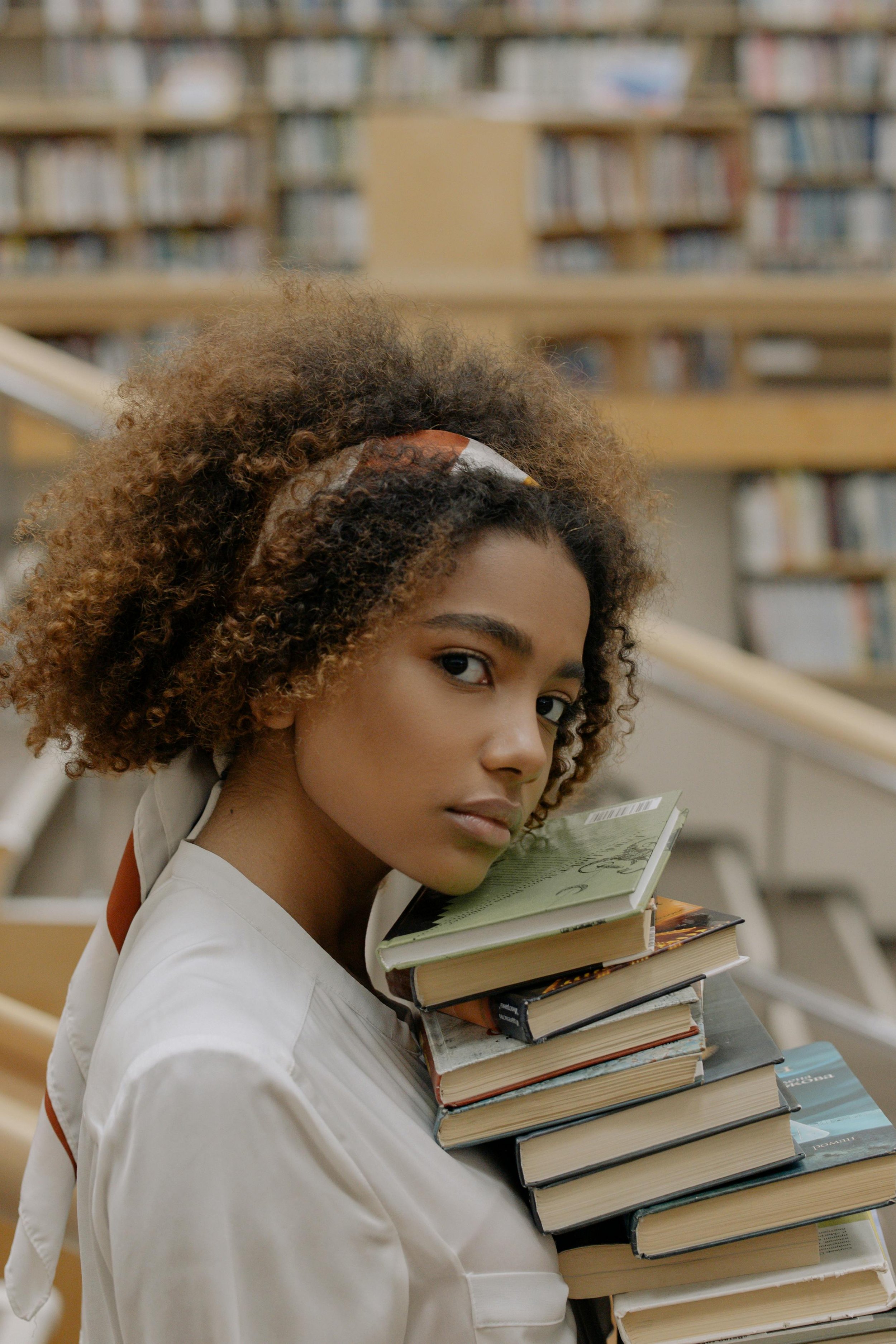 A young woman with curly hair wearing a white shirt and a headband, carrying a stack of books in a library