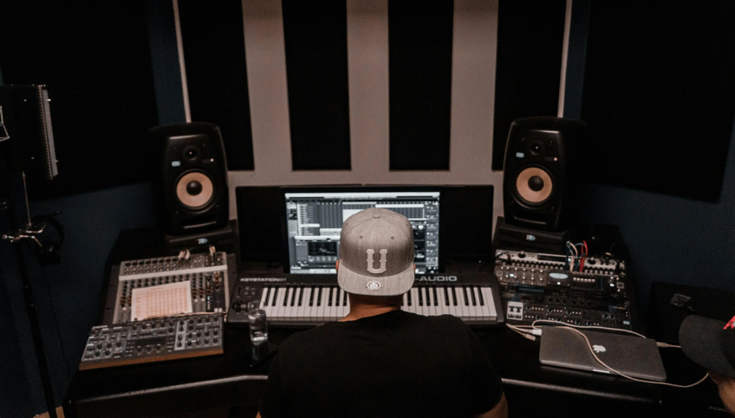 A person wearing a baseball cap with a university logo, sitting at a music production workstation in a dimly lit recording studio with acoustic panels and professional audio equipment.