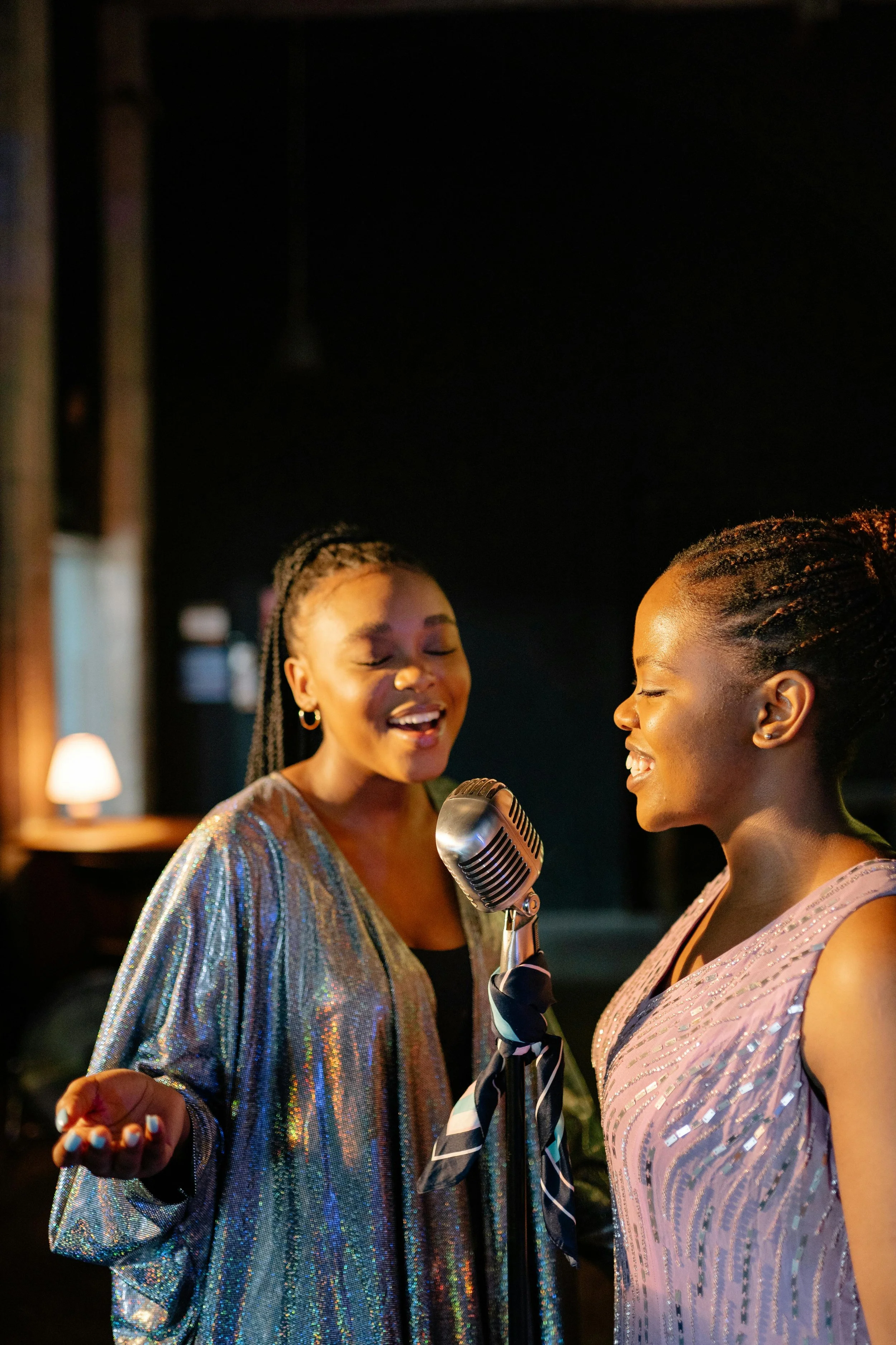 Two women singing into a microphone in a dimly lit room, both dressed in shiny, elegant outfits, enjoying the moment.