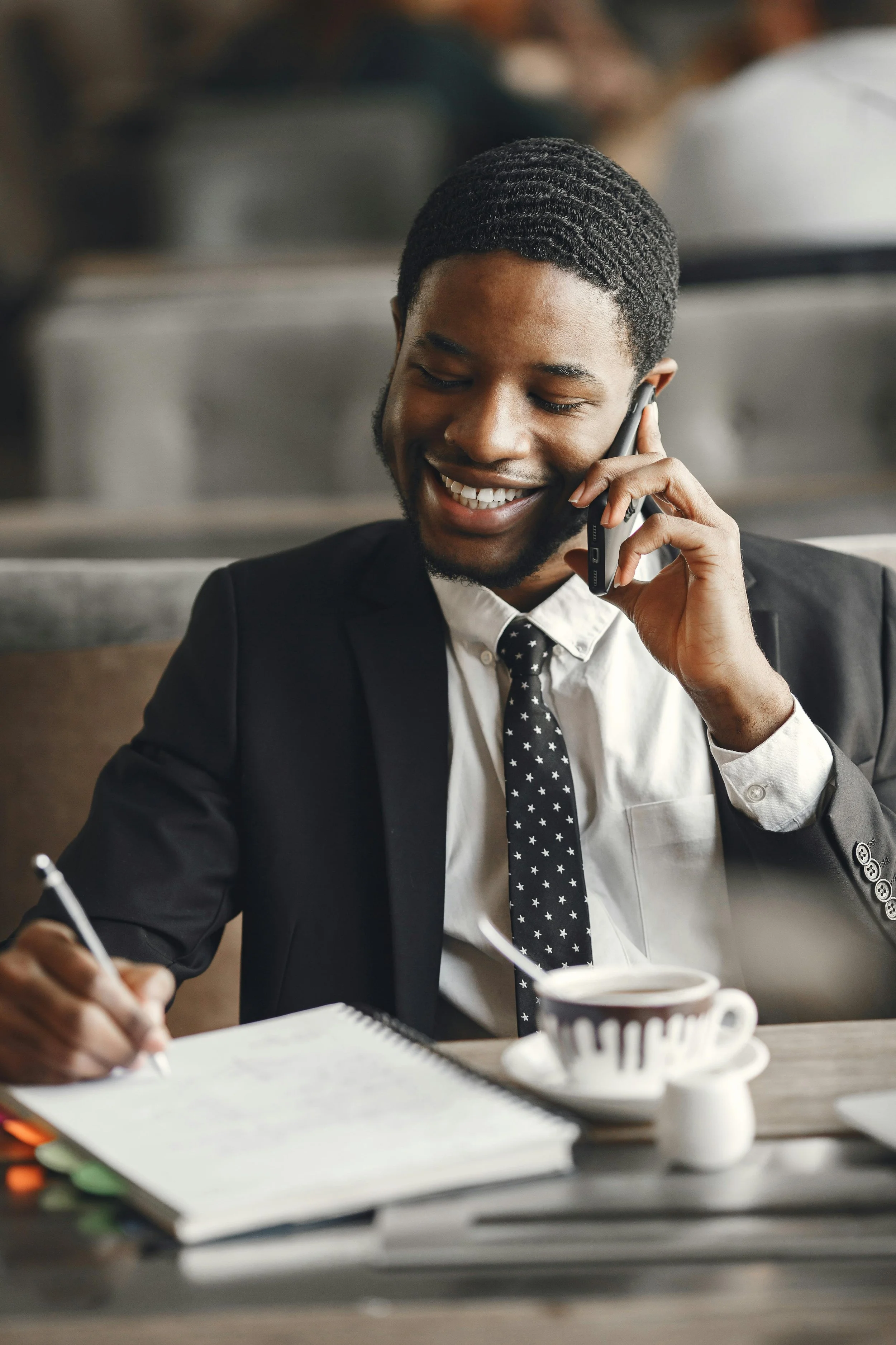 A young man in a business suit and tie, smiling while talking on a cell phone. He is sitting at a table with a notebook, a pen, and a coffee cup in a casual work environment.