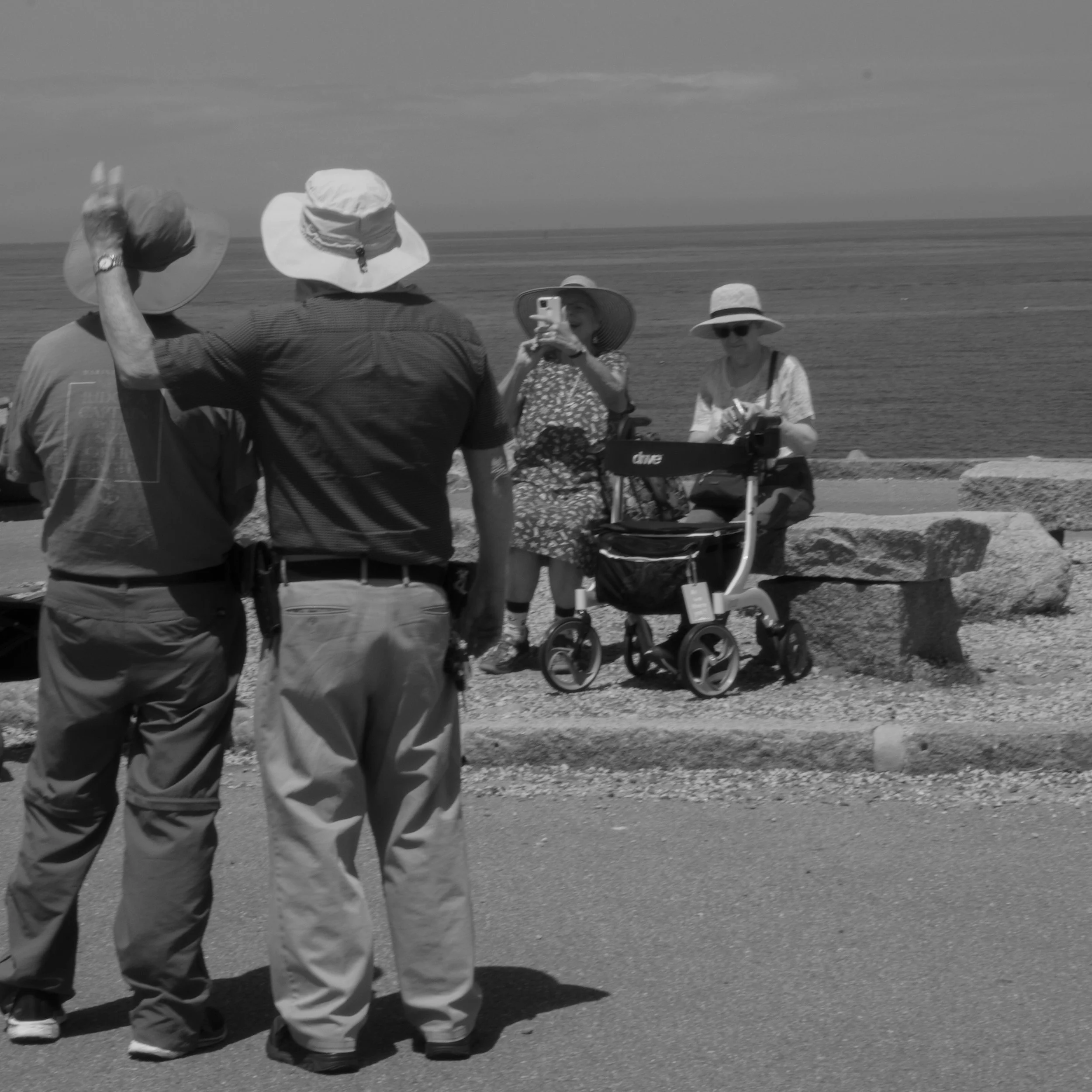 Four people stand near a body of water, with one person taking a photo of the others. They are wearing hats and casual clothing, and there's a stroller nearby. The scene appears to be at a beach or lakeside.